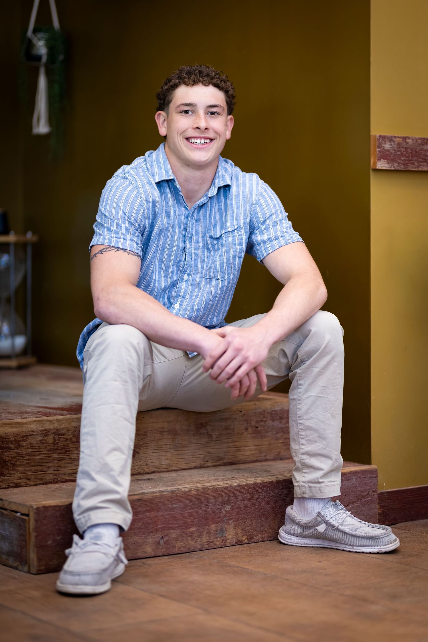 Smiling young man in blue striped shirt and khaki pants sitting relaxed on wooden steps indoors.