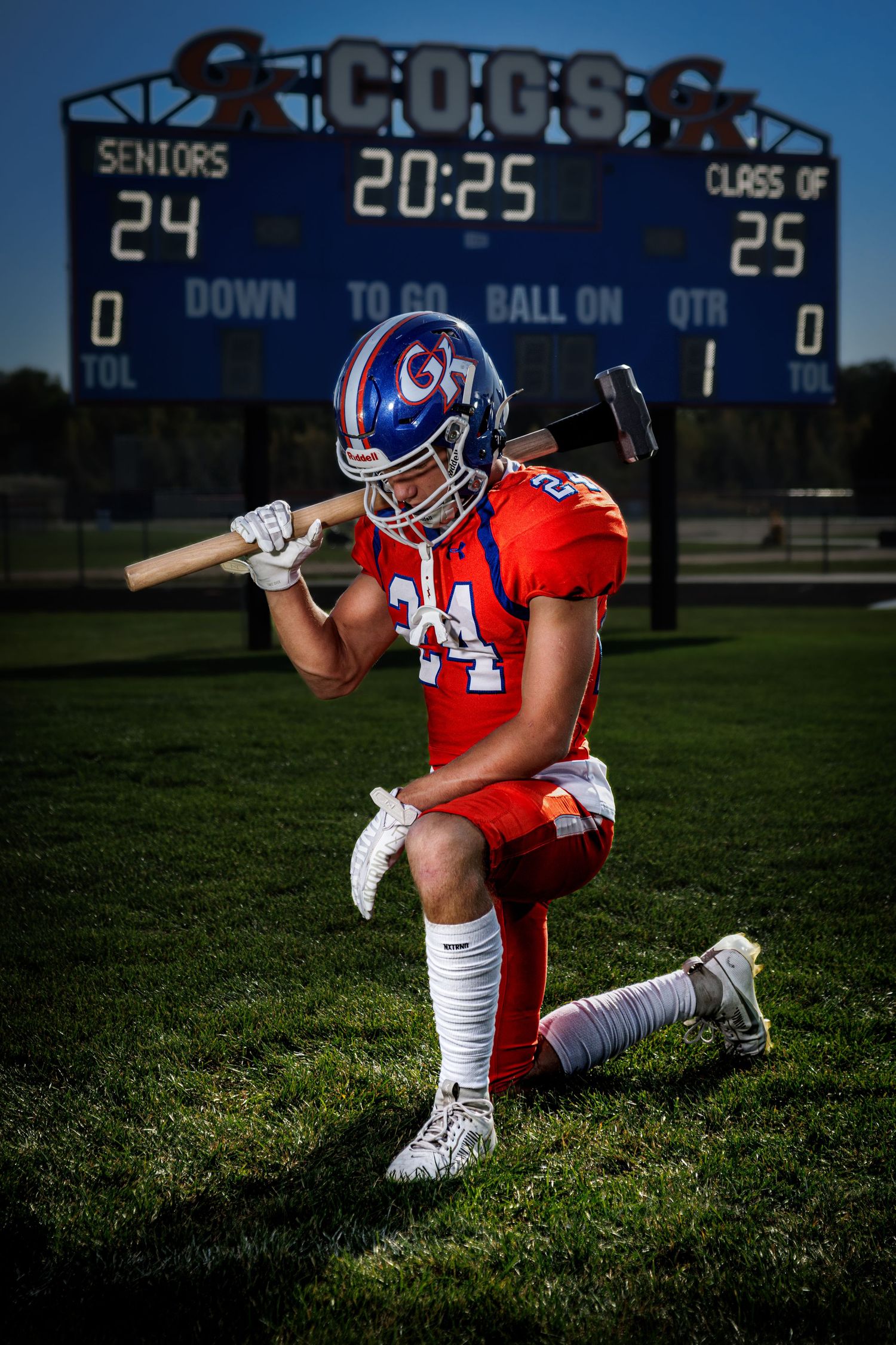 Football player in red uniform kneeling on field holding a sledgehammer with COGS scoreboard behind him.