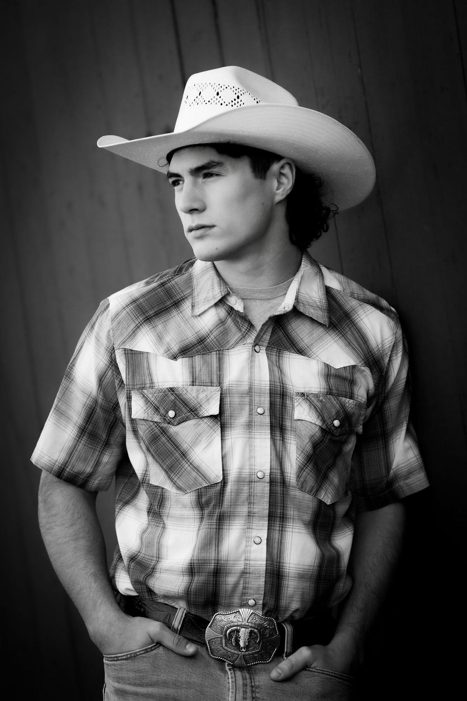 Young man in cowboy hat and plaid western shirt with belt buckle poses in black and white portrait.