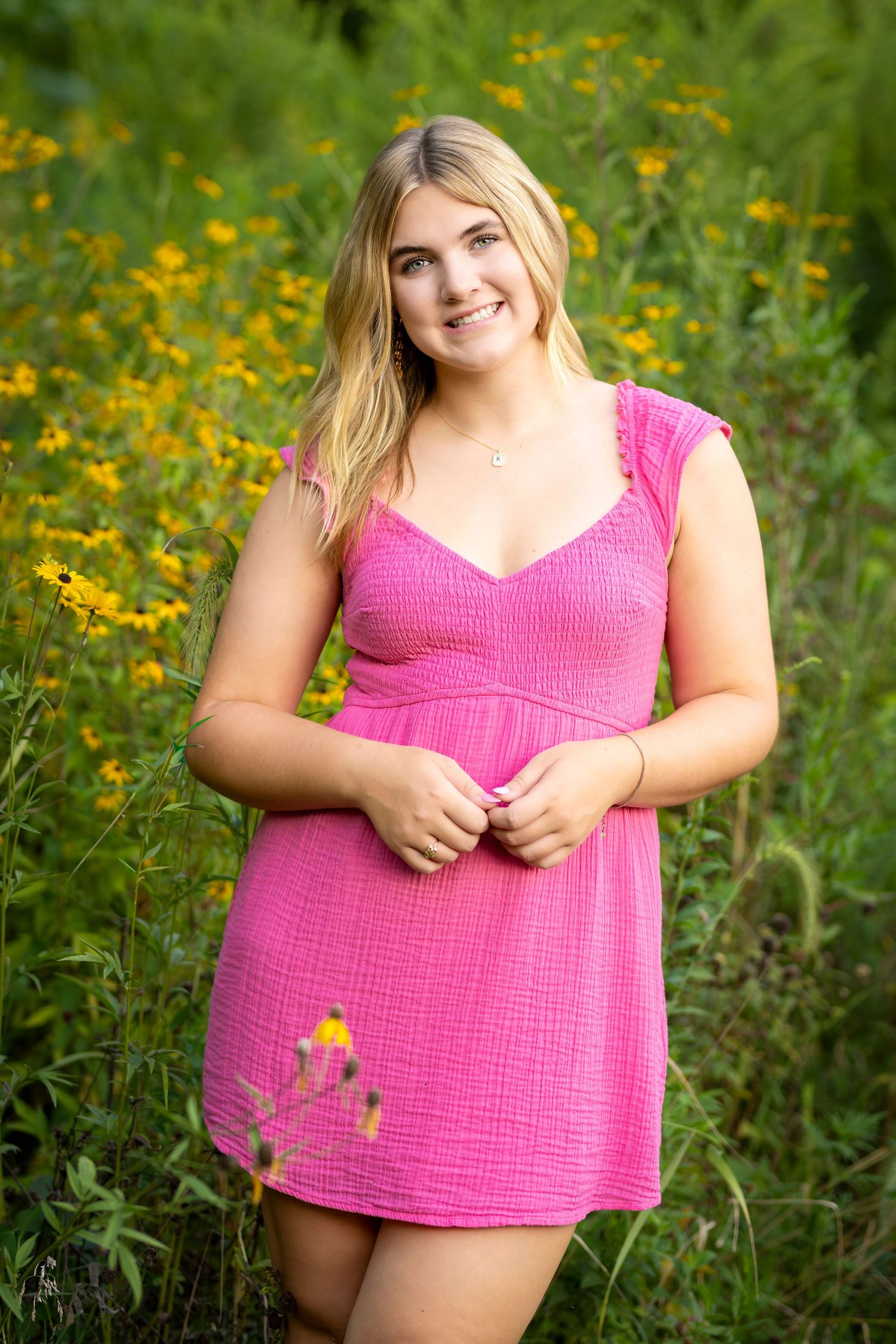 Young blonde woman in pink dress smiling among yellow wildflowers in a garden.