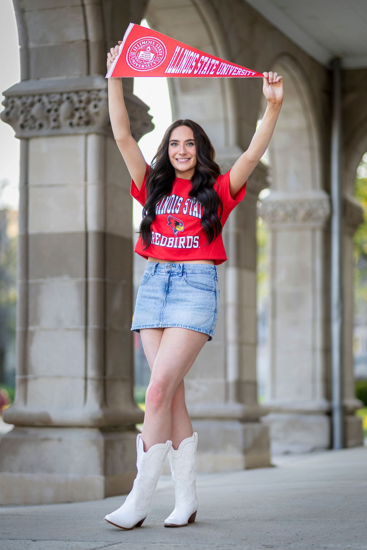 Young woman in Illinois State University Redbirds shirt and denim skirt holds pennant while wearing white cowboy boots.