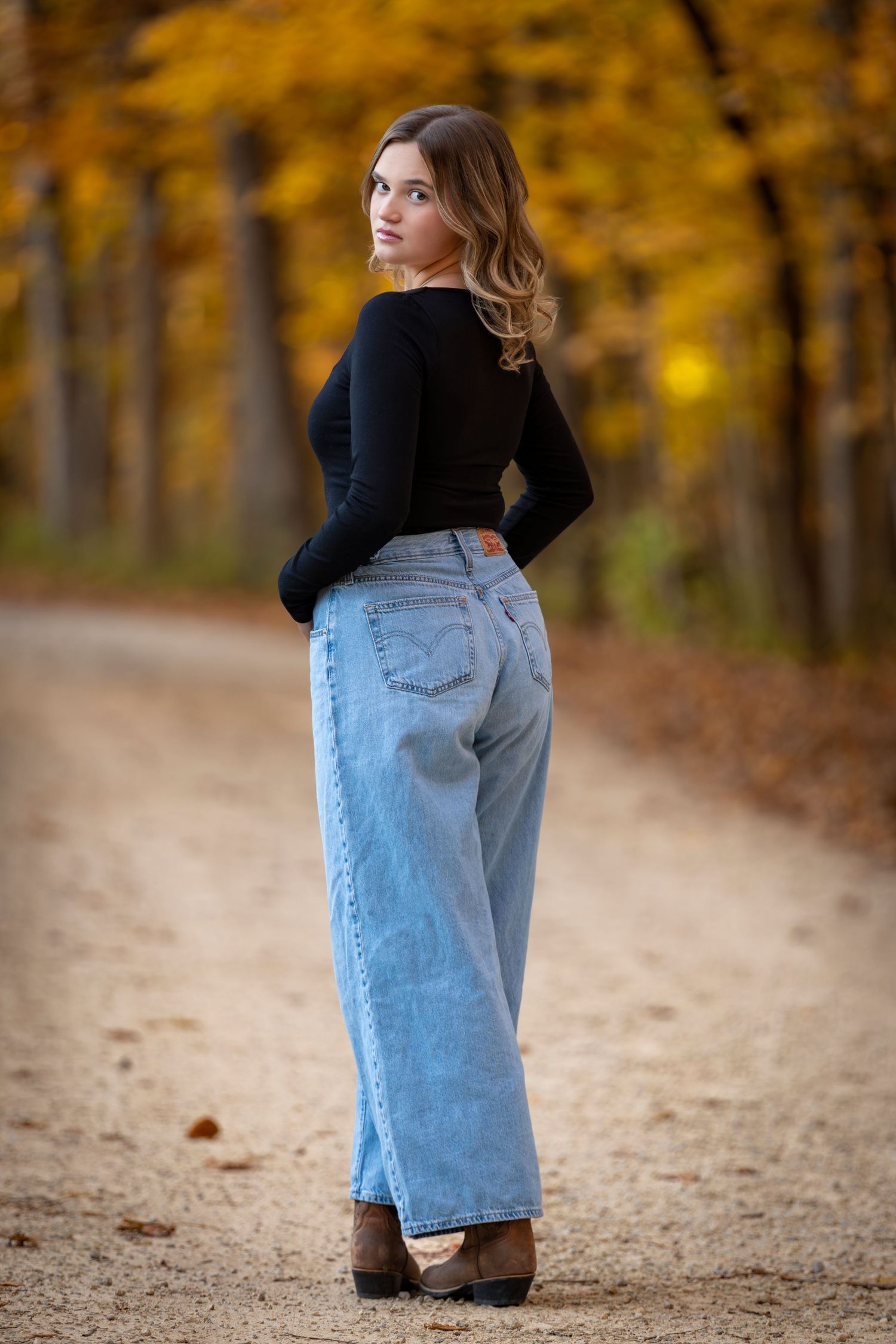 Young woman in wide-leg jeans and black top poses on autumn forest path surrounded by golden foliage.