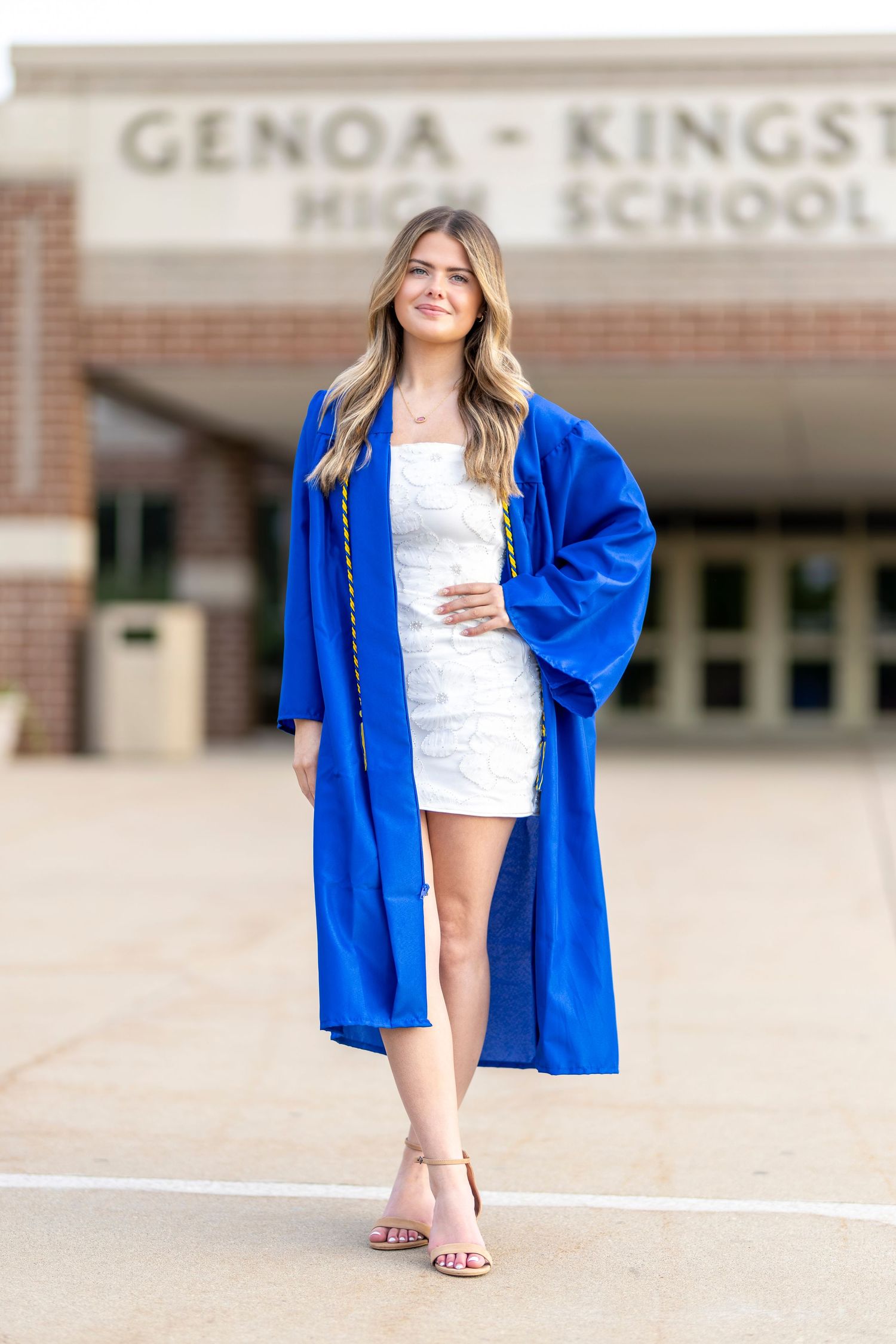 A smiling graduate in a royal blue cap and gown poses in front of Genoa-Kingston High School wearing a white dress.