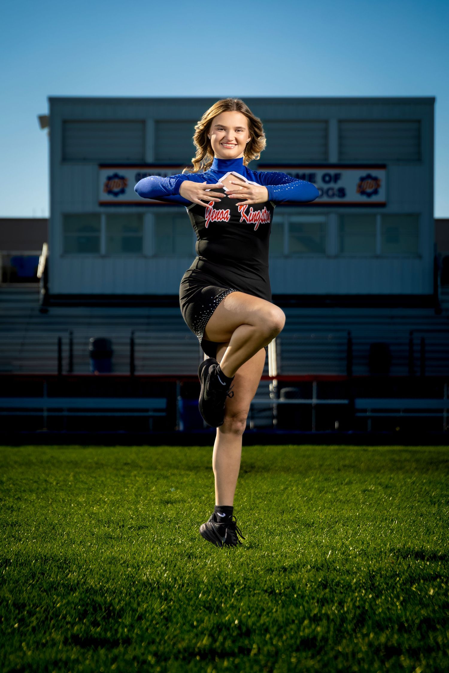 Cheerleader in blue and black uniform poses on green field with stadium bleachers in background on sunny day.