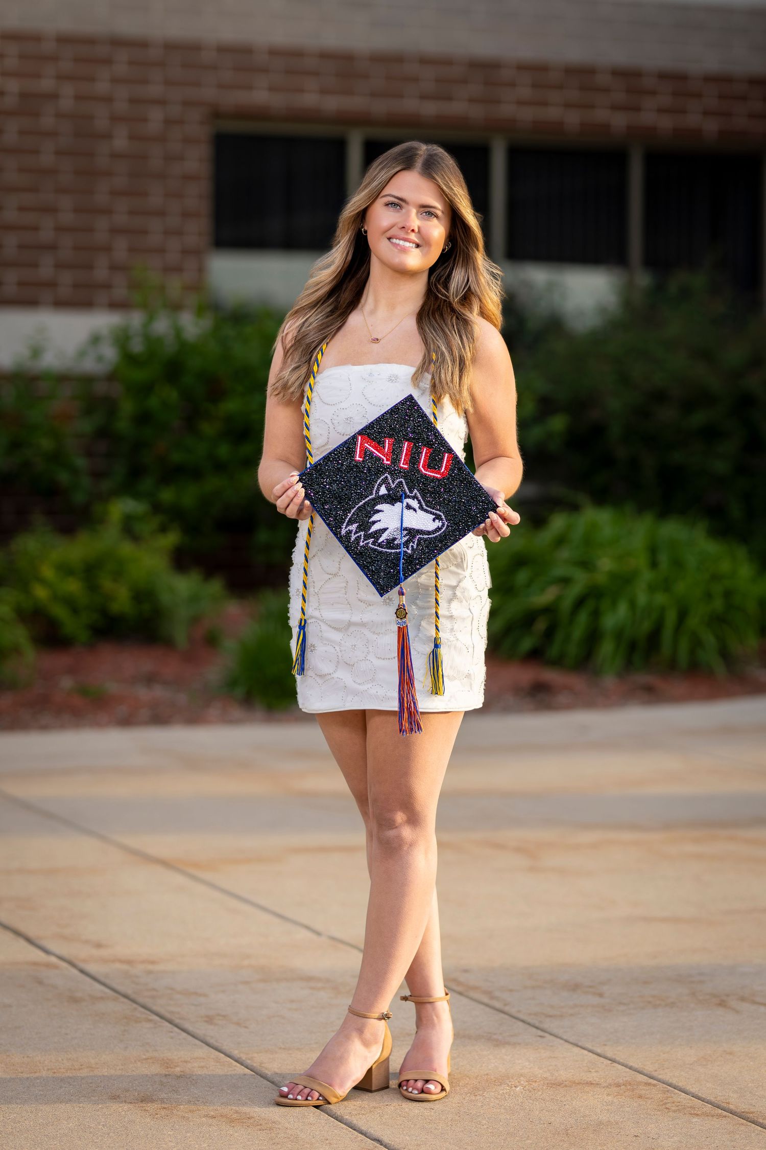 A smiling young woman in a white dress holds an NIU graduation cap outdoors near a brick building.