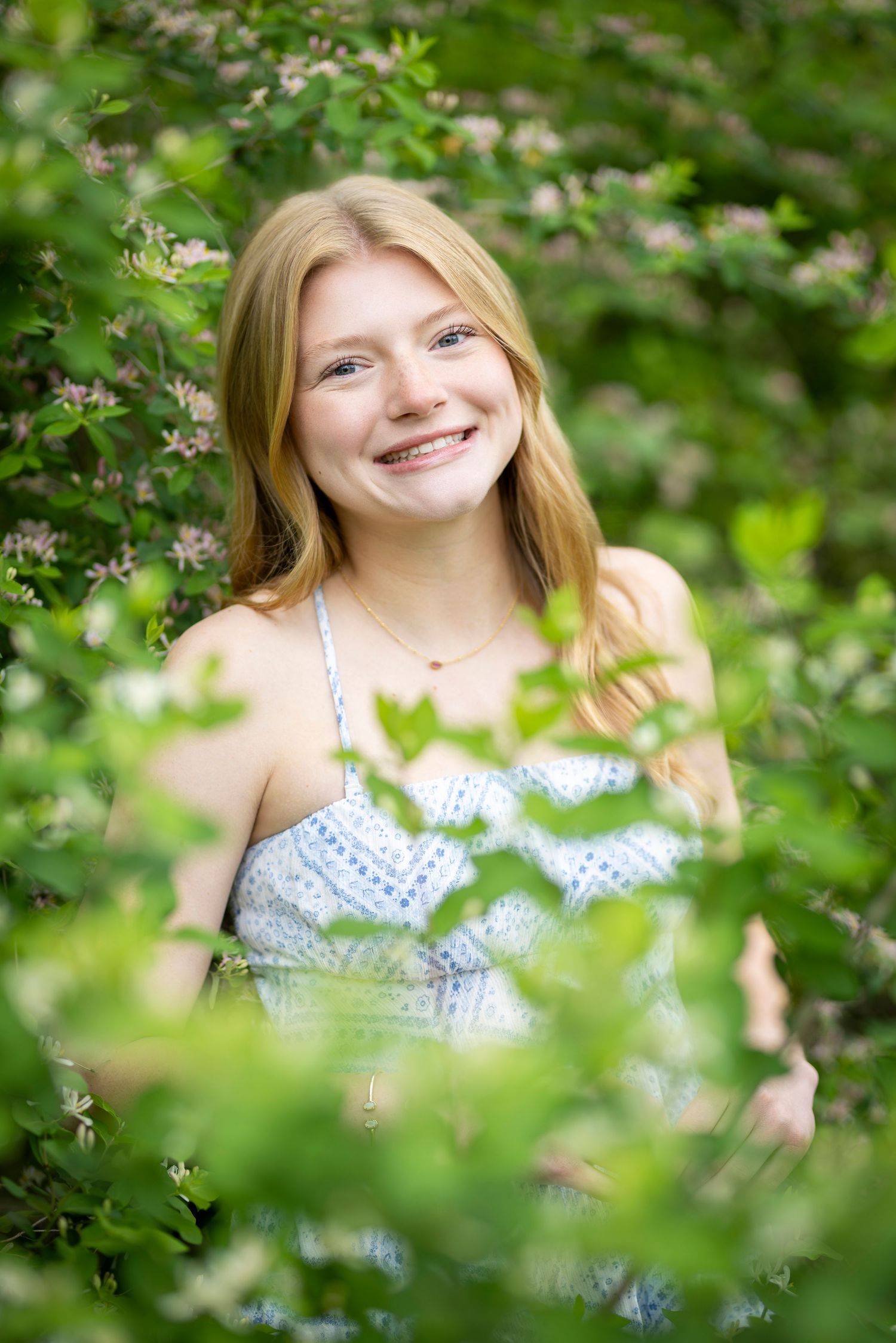 Smiling teenage girl with long blonde hair wearing a blue sundress, surrounded by lush green foliage and pink flowers.