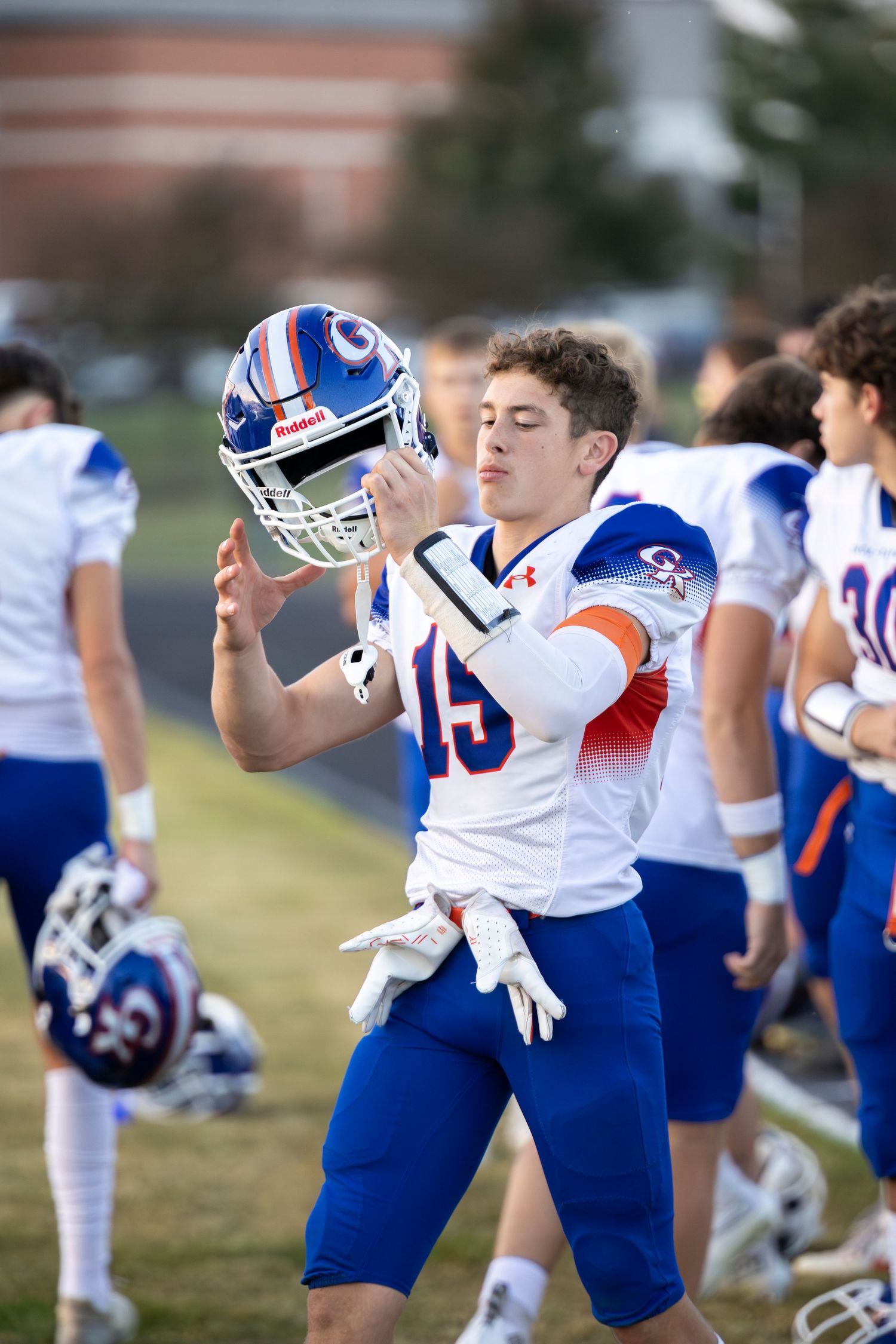 High school football player in white and blue uniform holding helmet on sideline during game.