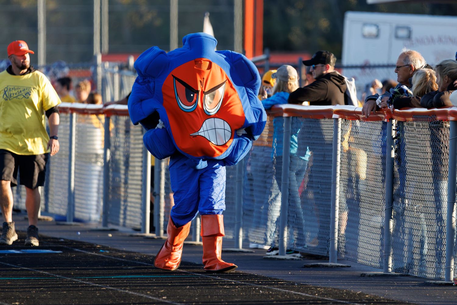 A mascot in orange and blue costume walks along a fenced track at an outdoor sporting event.