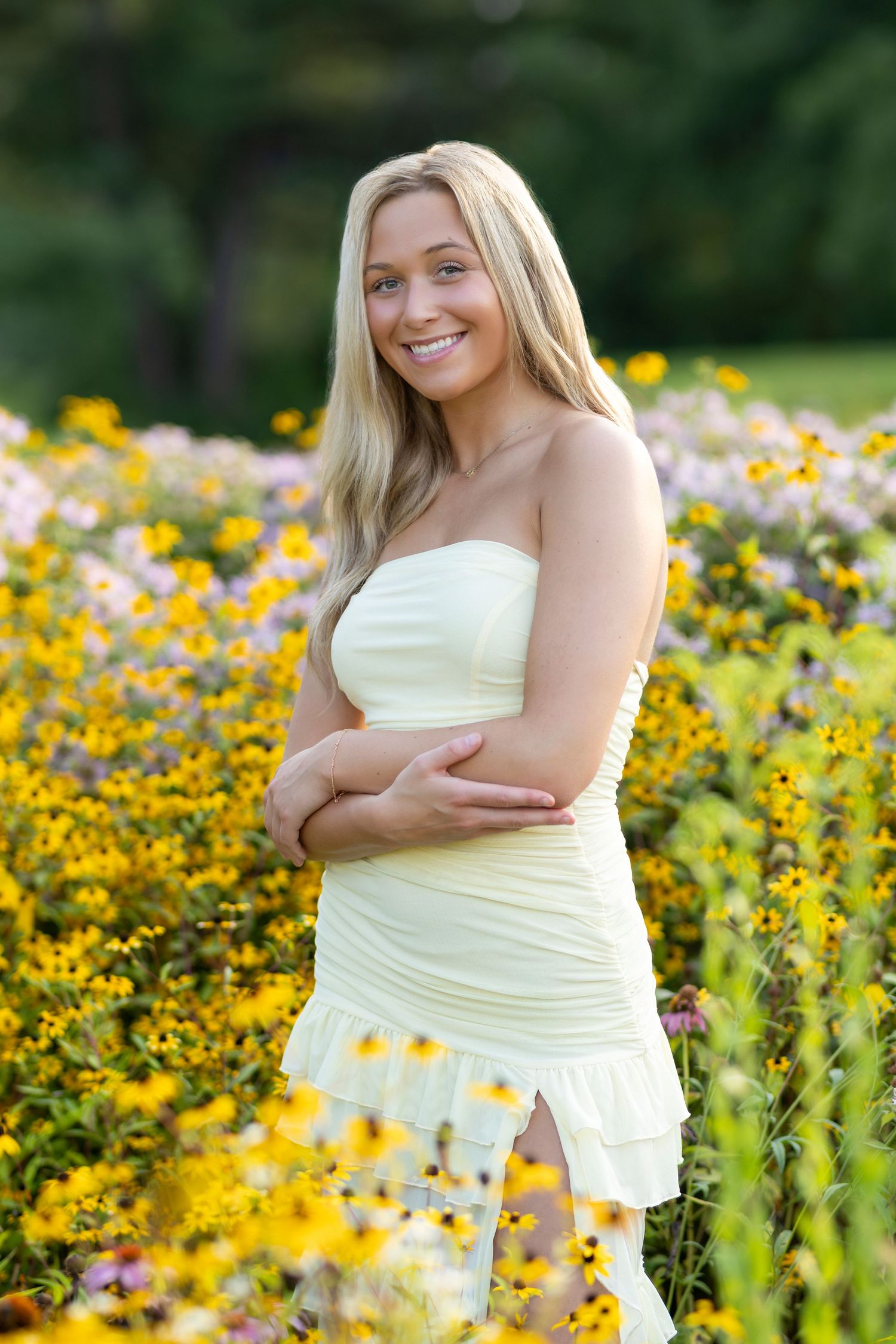Smiling blonde woman in a white strapless dress posing in a vibrant garden of yellow and pink flowers.