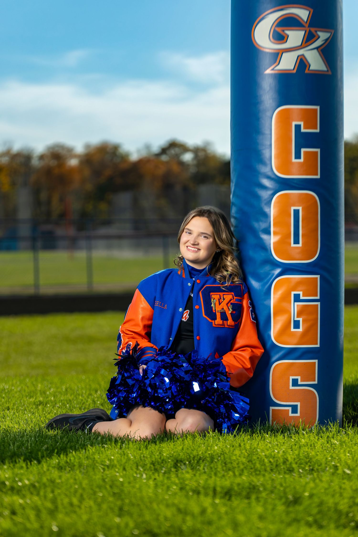 A cheerleader in a GK Cogs letterman jacket sits on grass holding blue pom-poms beside a blue team goal post pad.