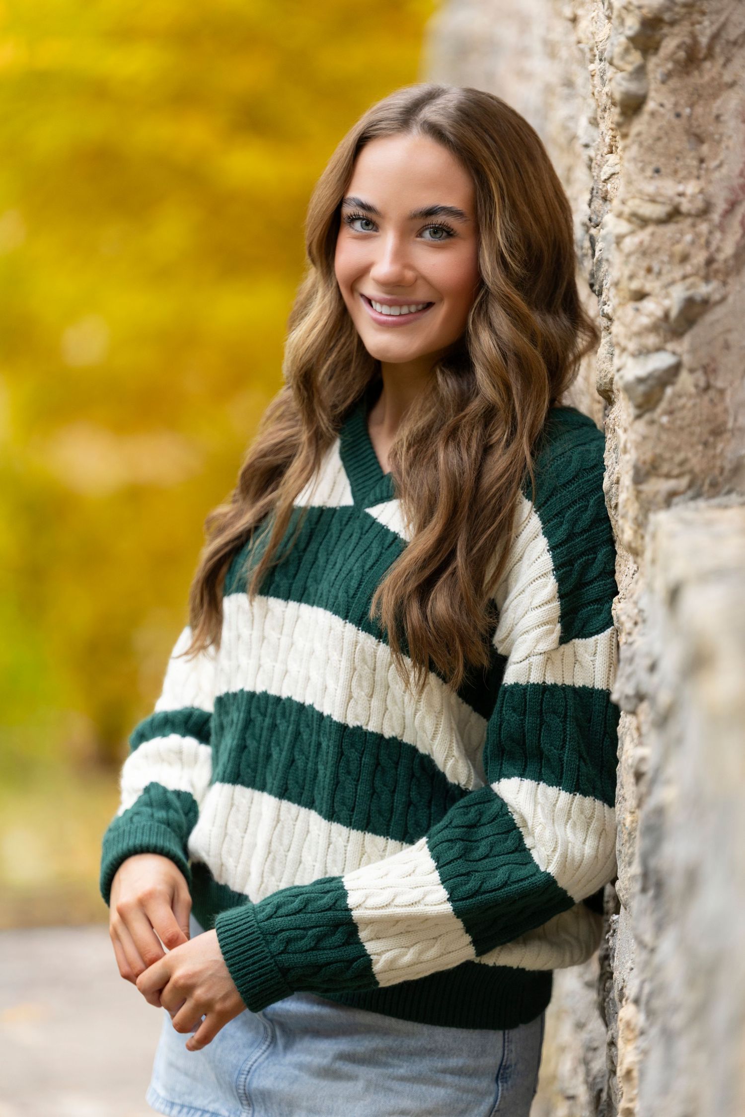 Young woman in green and white cable-knit striped sweater smiling outdoors against stone wall with autumn foliage.