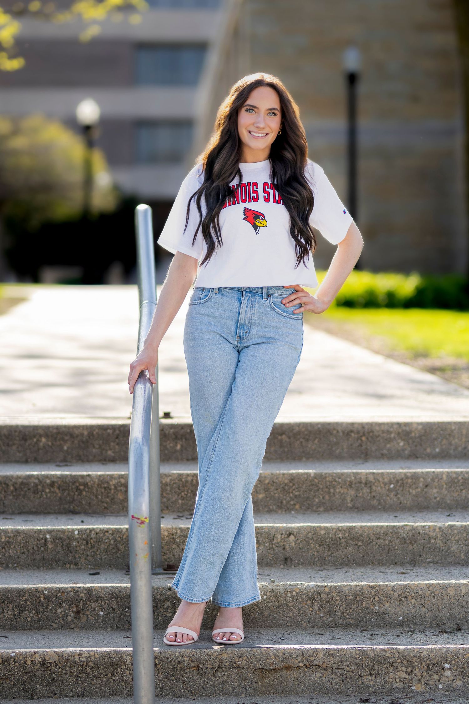 Young woman in Illinois State University crop top and jeans posing on outdoor steps on a sunny day.
