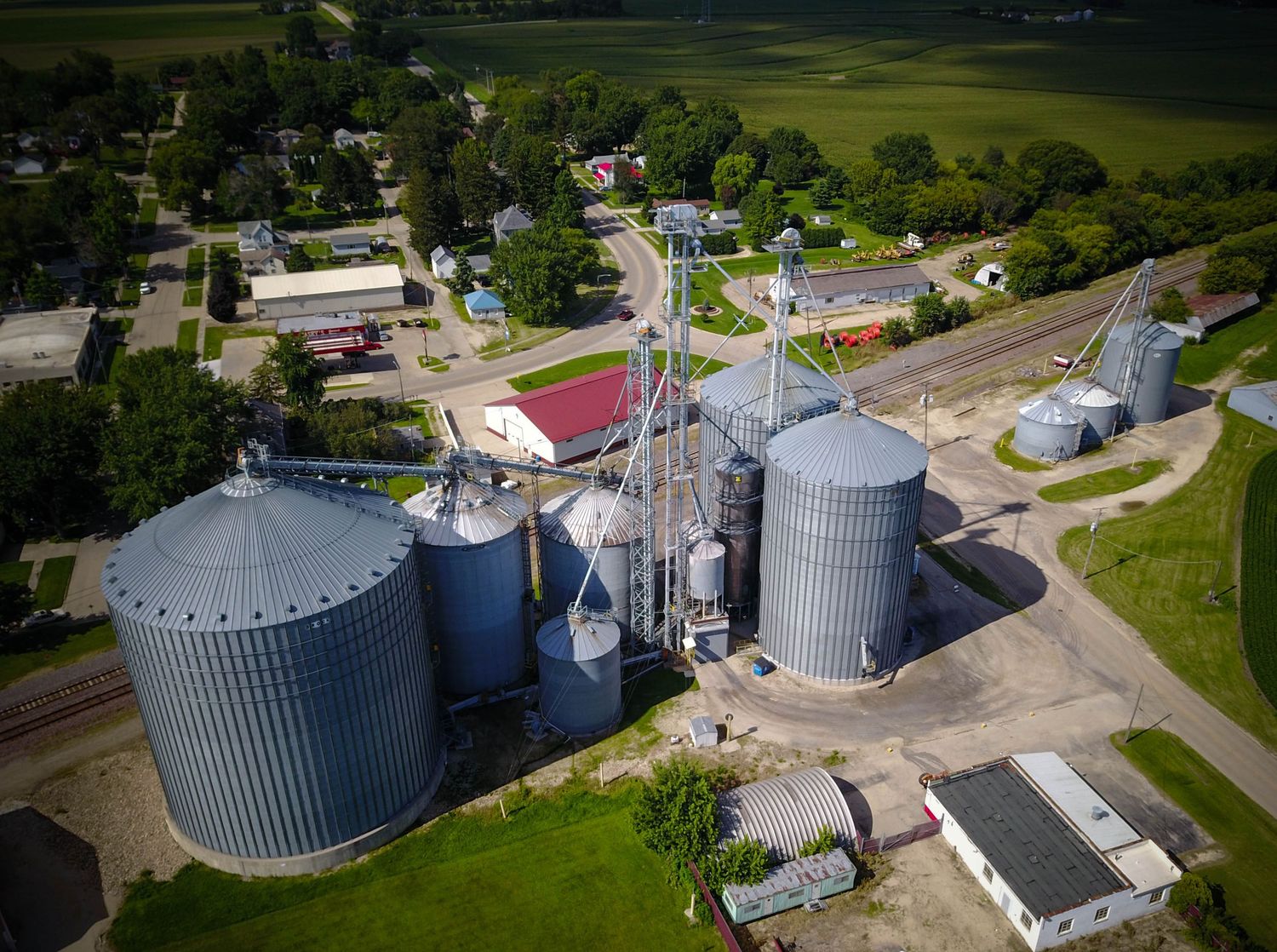 Aerial view of large grain storage silos at a rural farm elevator near a small town with railroad tracks.