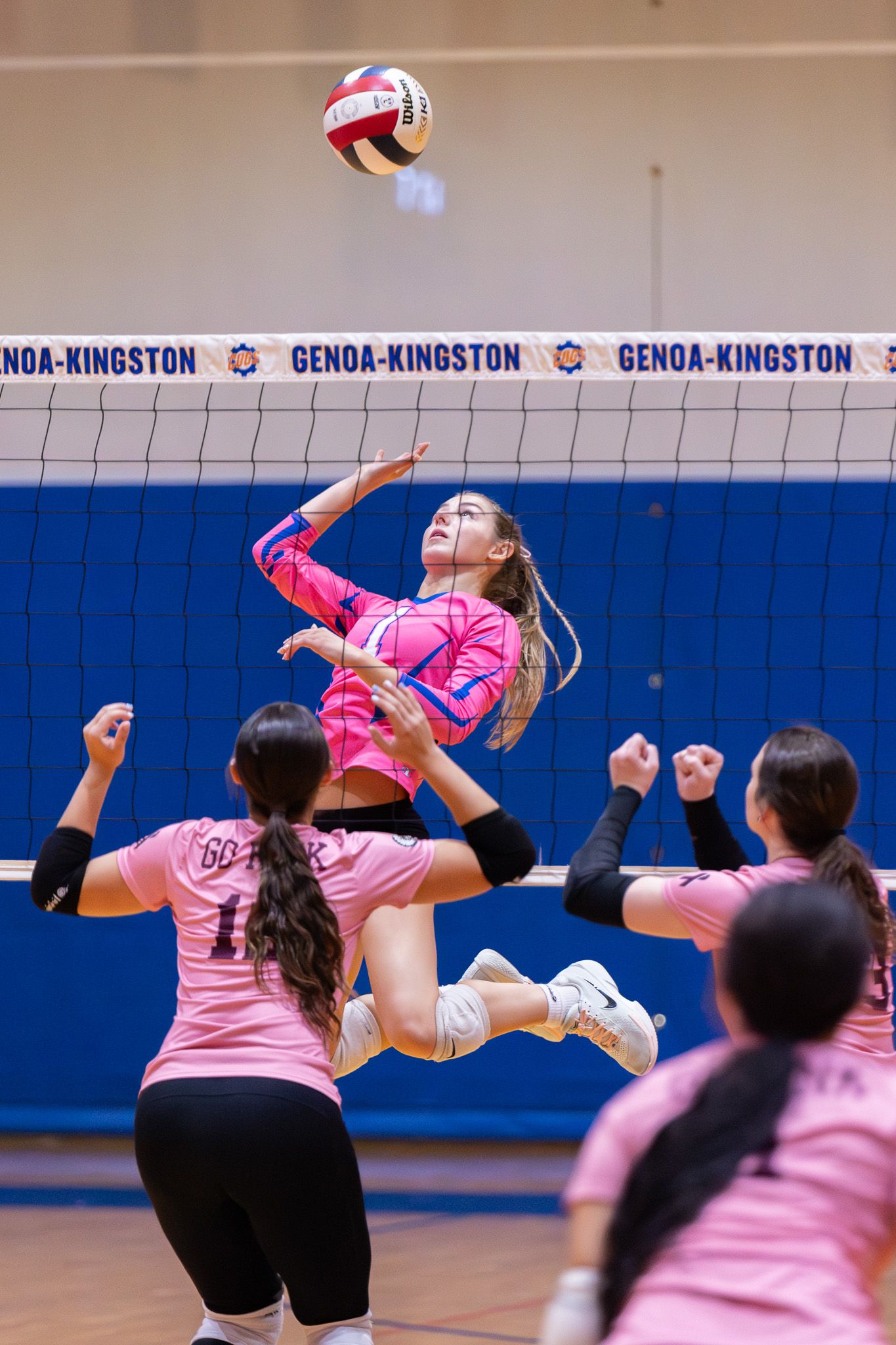 Female volleyball player in pink uniform jumps to spike ball over net at Genoa-Kingston gym.