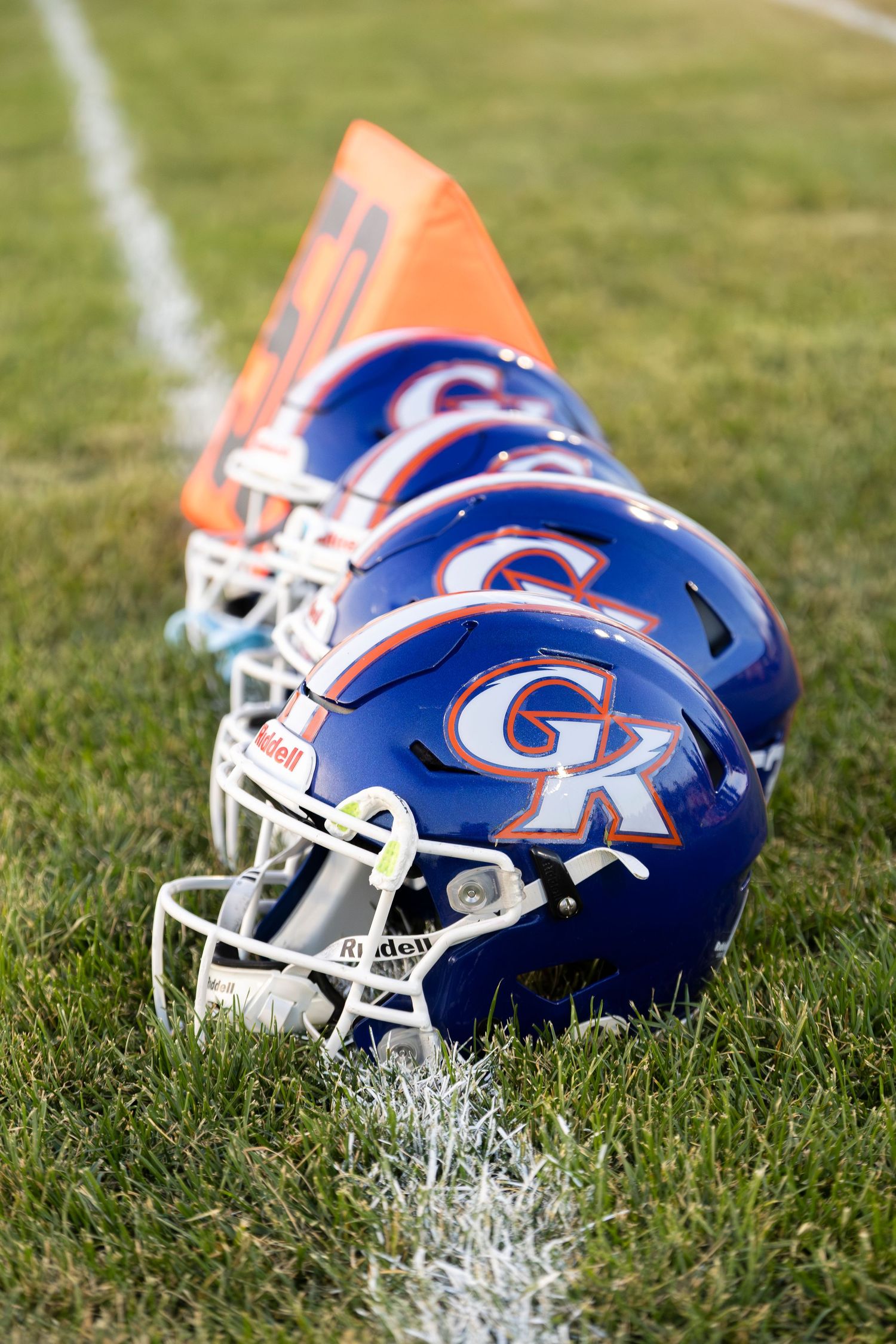 Blue GK football helmets lined up on grass field next to orange yard marker.