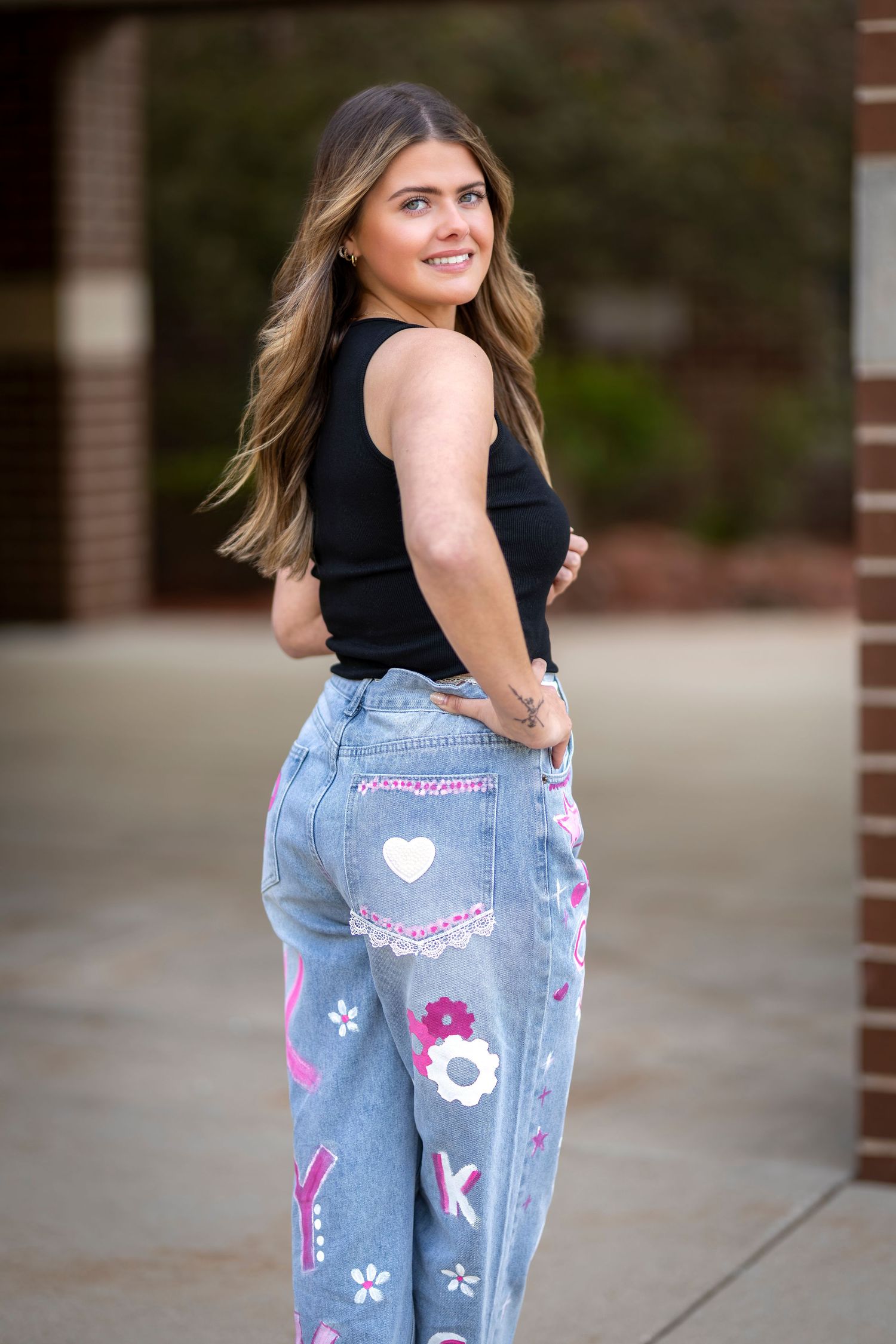 Young woman smiling in decorated pink denim jeans and black top, posing outdoors near brick building.