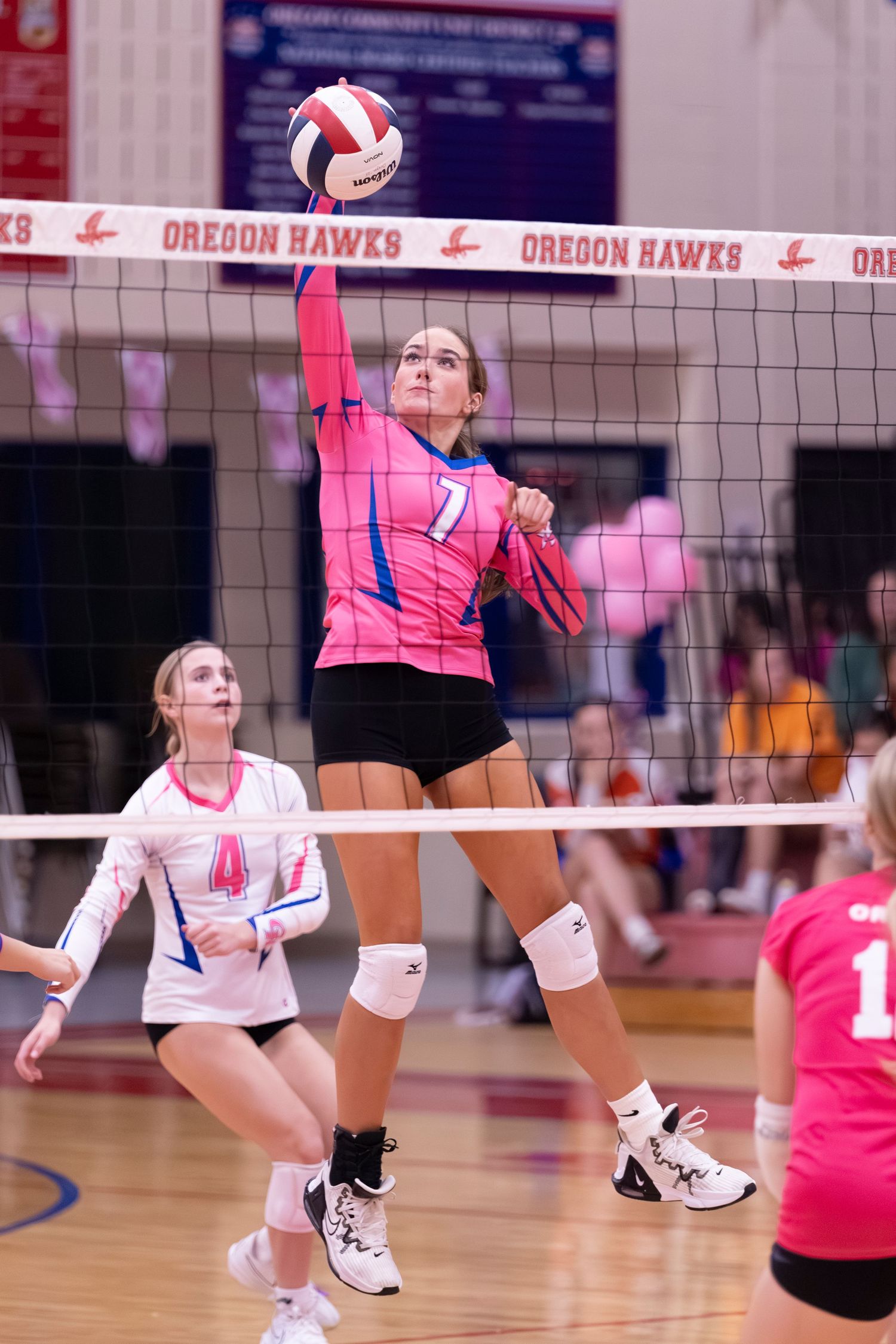 Volleyball player #7 in pink jersey jumps to spike the ball over the net during a competitive match.