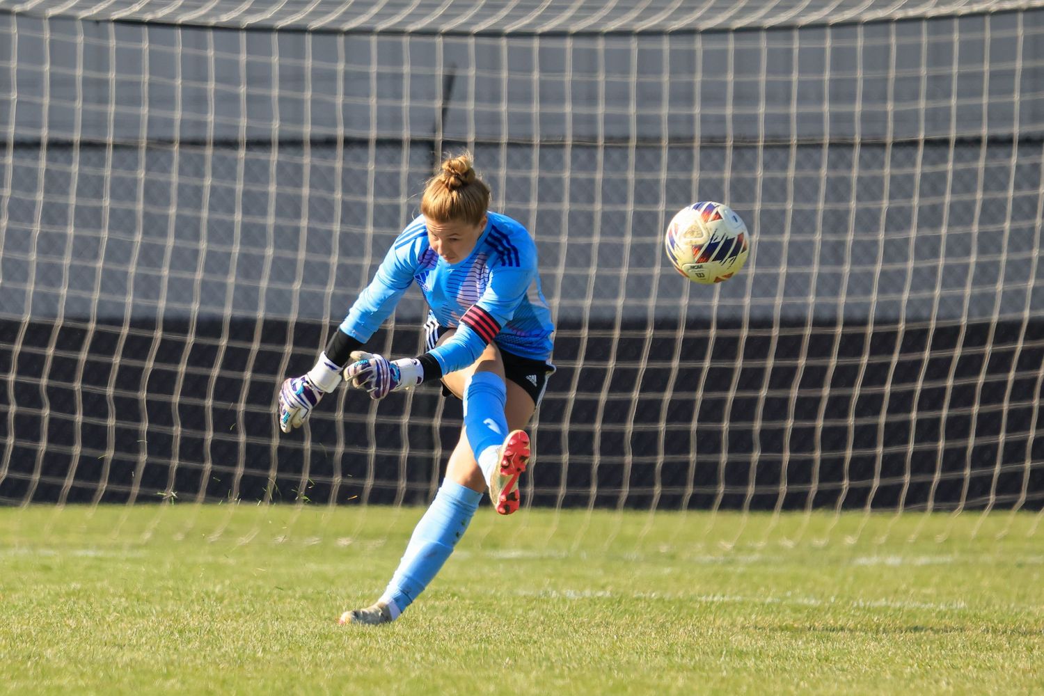 Female soccer goalkeeper in blue uniform kicking ball during a game near the net on a sunny day.