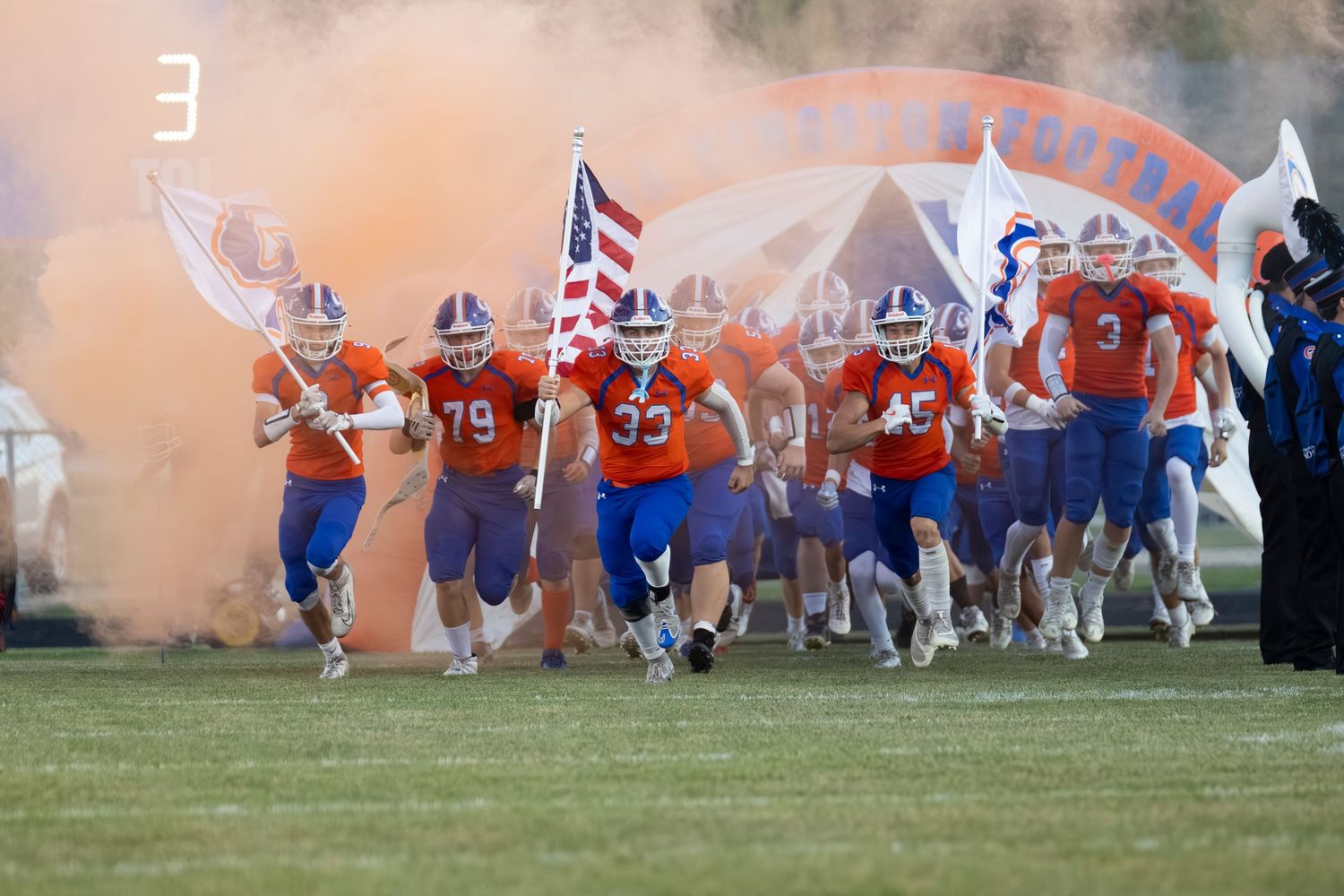 Football team in orange uniforms runs onto field with American flag and orange smoke during pre-game entrance.
