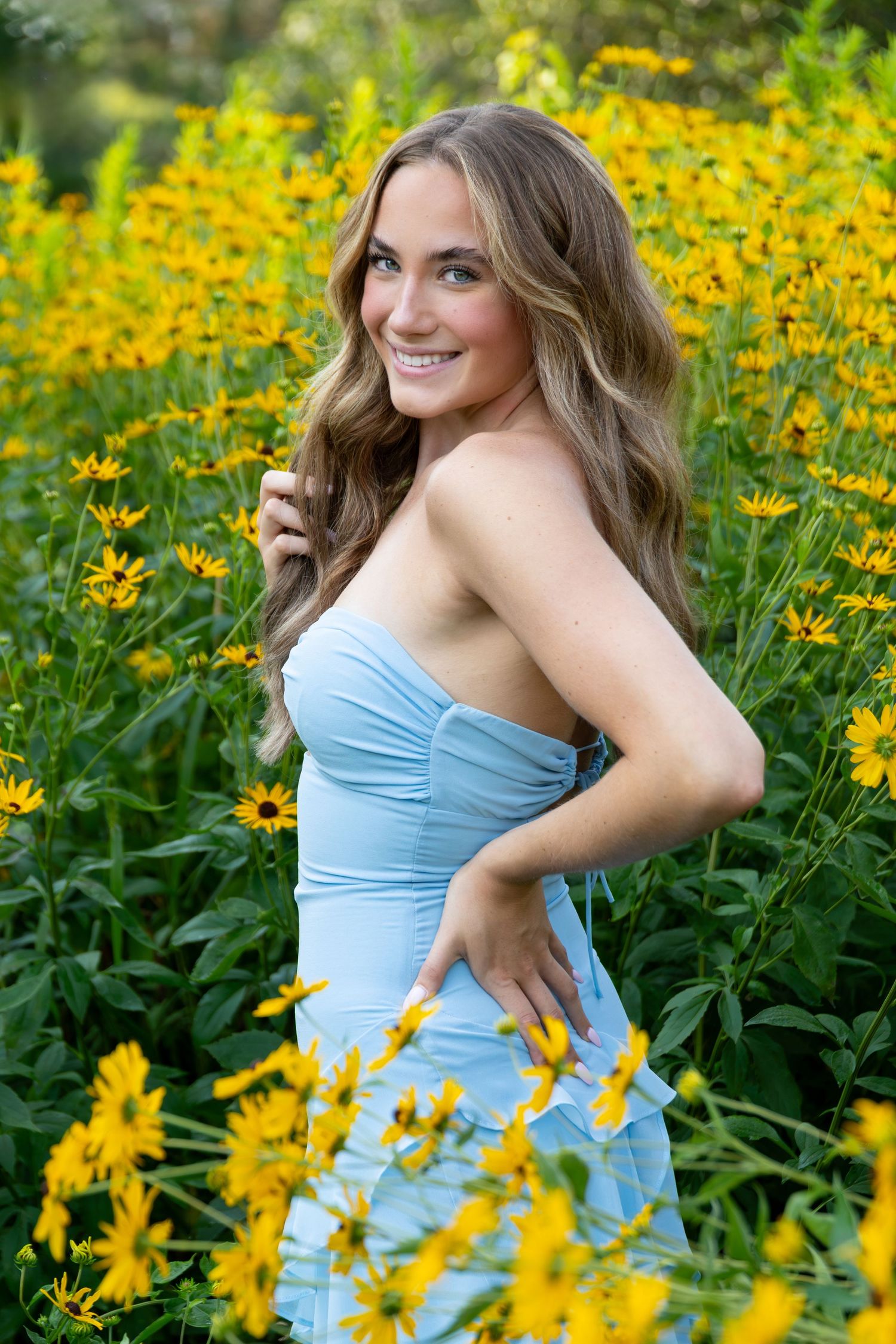 Young woman in light blue strapless dress smiling among vibrant yellow wildflowers in a lush garden setting.