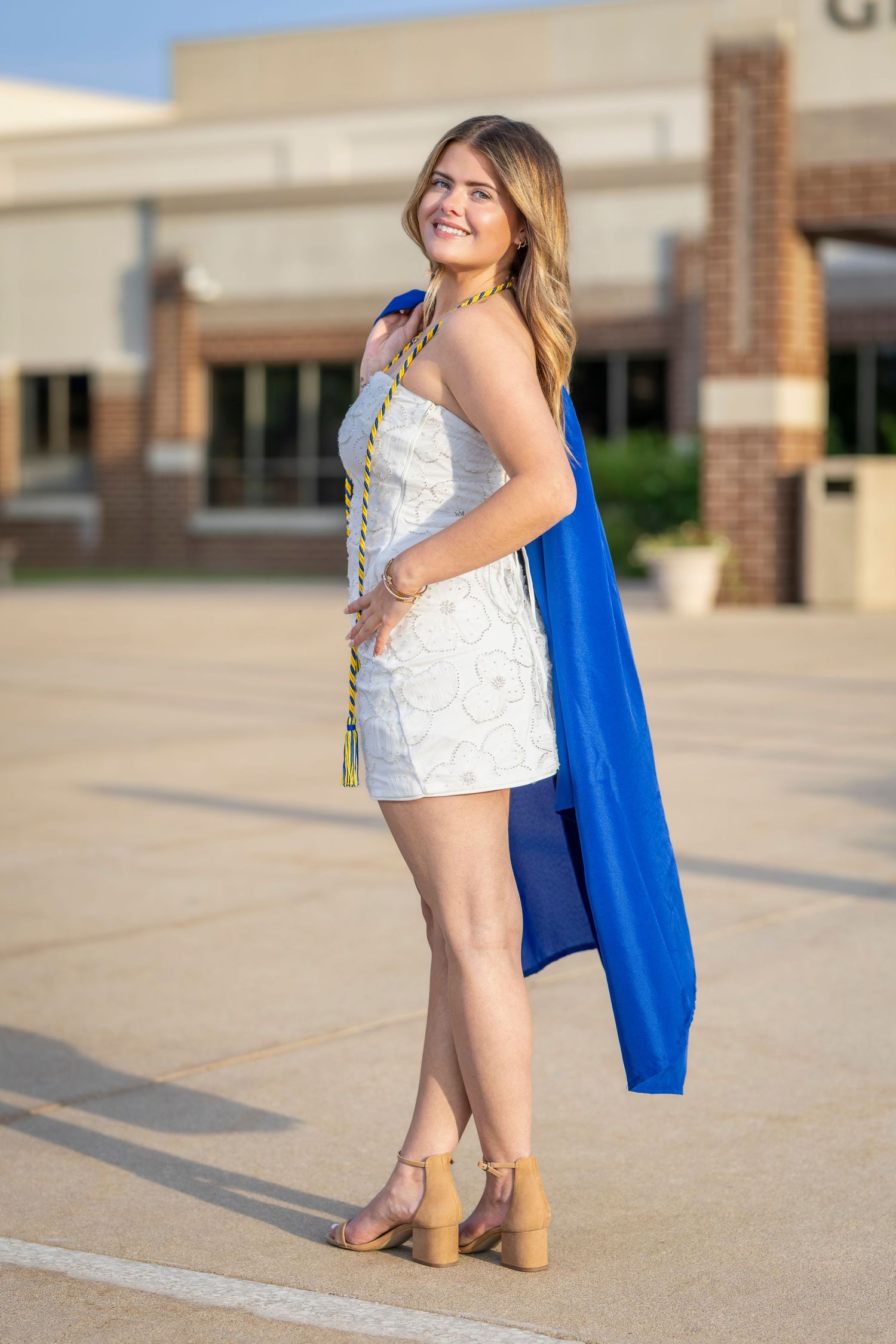 Smiling graduate in white dress holding blue graduation gown and gold honor cords outside school building.
