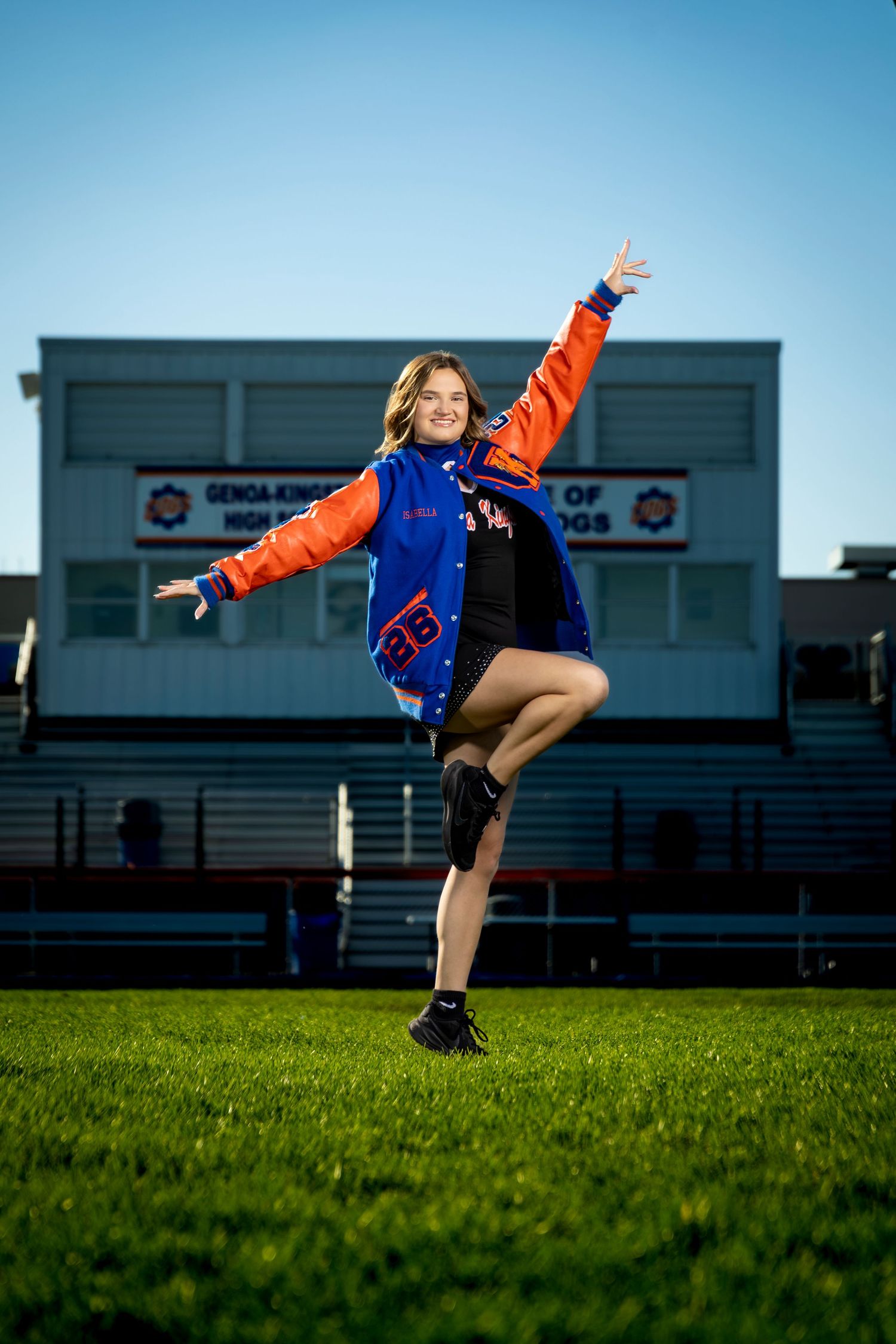 A cheerful teen in a blue and orange varsity jacket jumps joyfully on a green football field at a high school stadium.