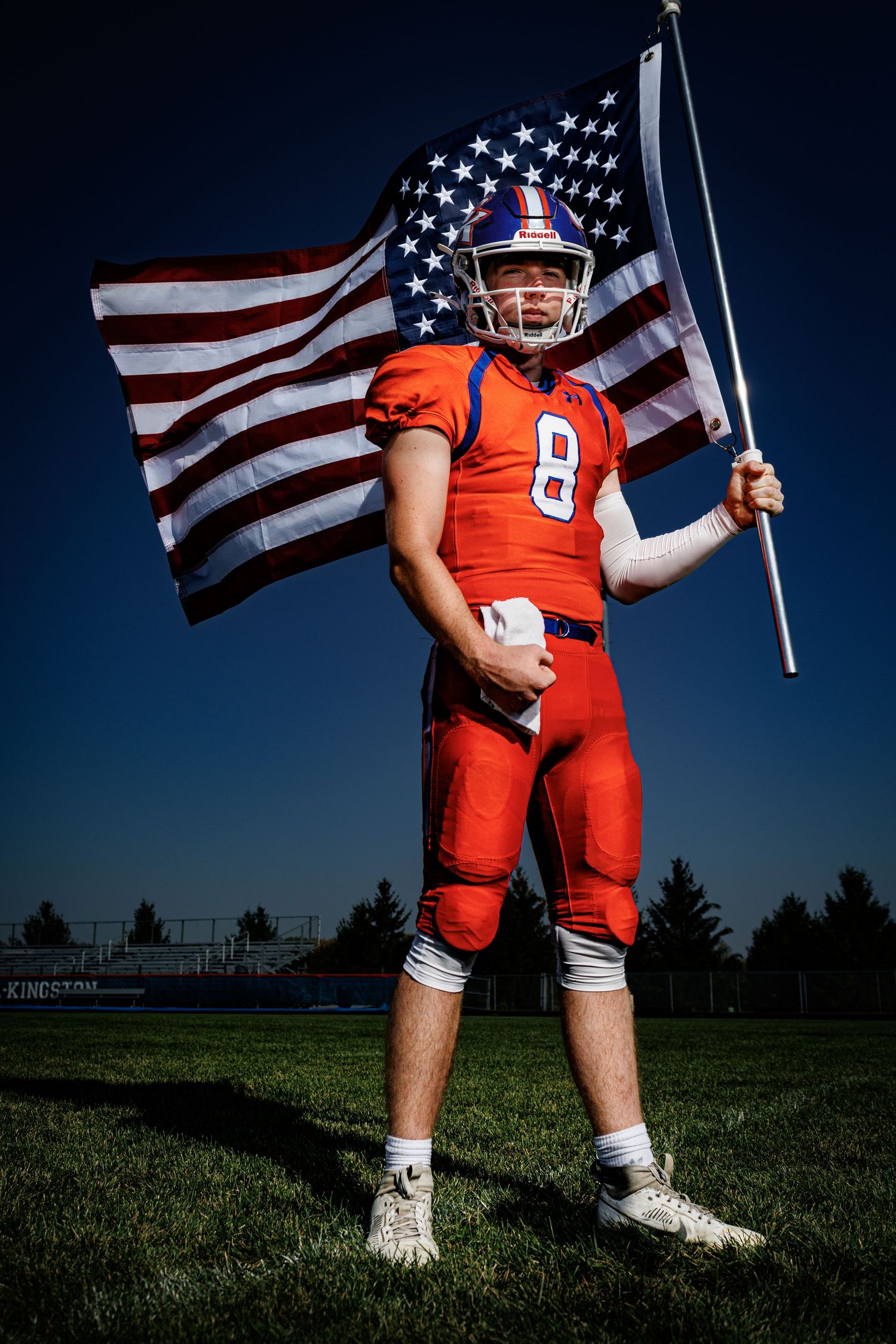 Football player #8 in red uniform holds American flag on field under blue sky.