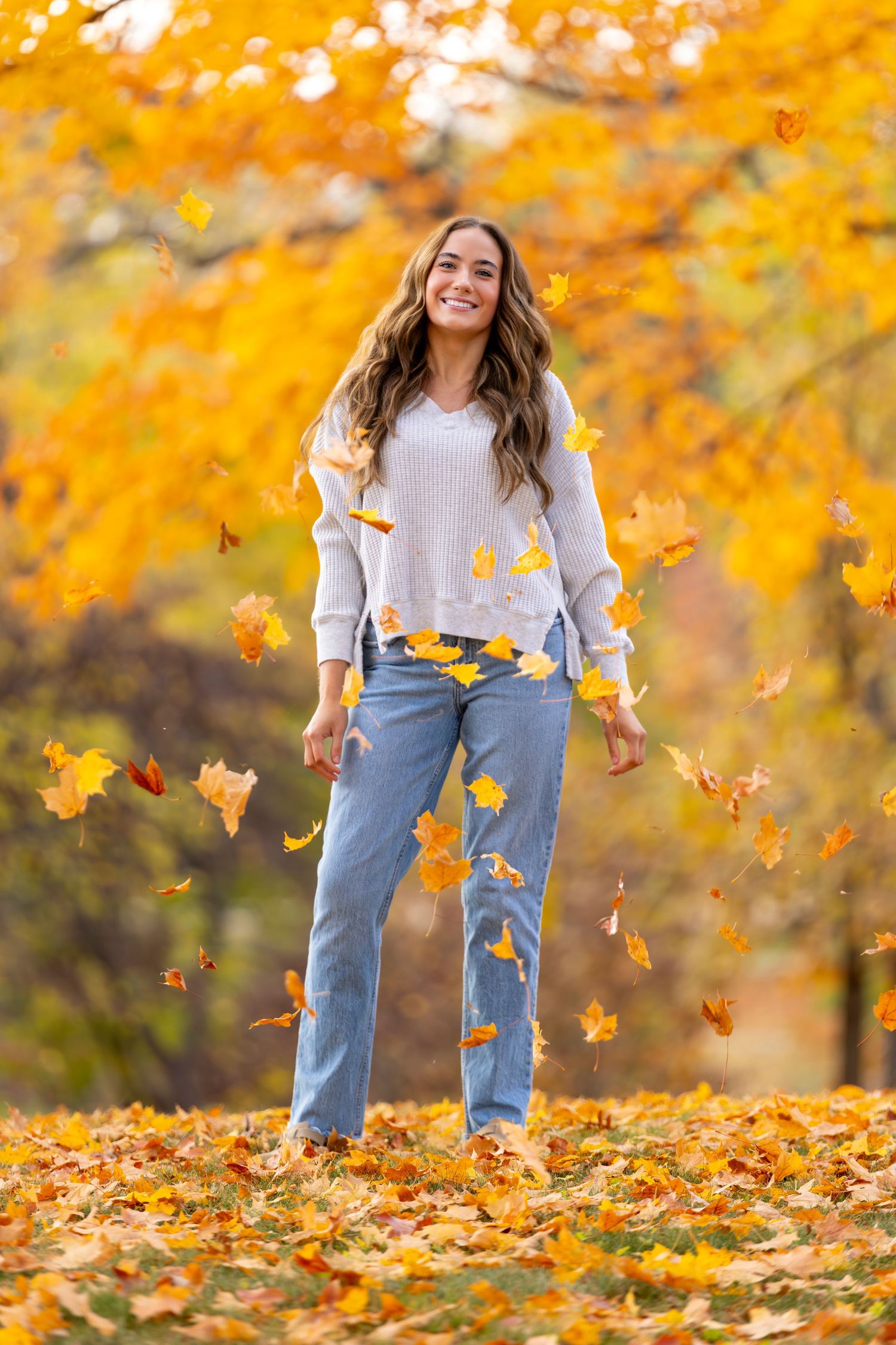 Woman smiling in autumn park with falling yellow maple leaves, wearing white sweater and jeans.