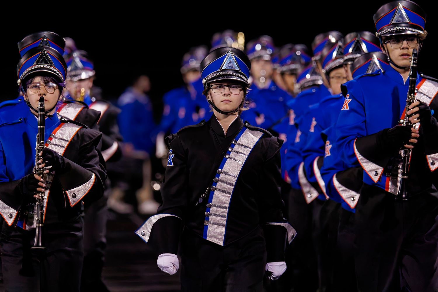 Marching band members in blue and black uniforms perform on a dark field during a nighttime competition.