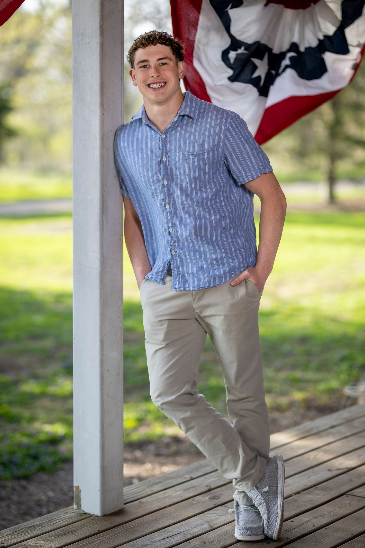 Smiling young man in blue striped shirt and khakis leaning against white pillar with American flag backdrop outdoors.
