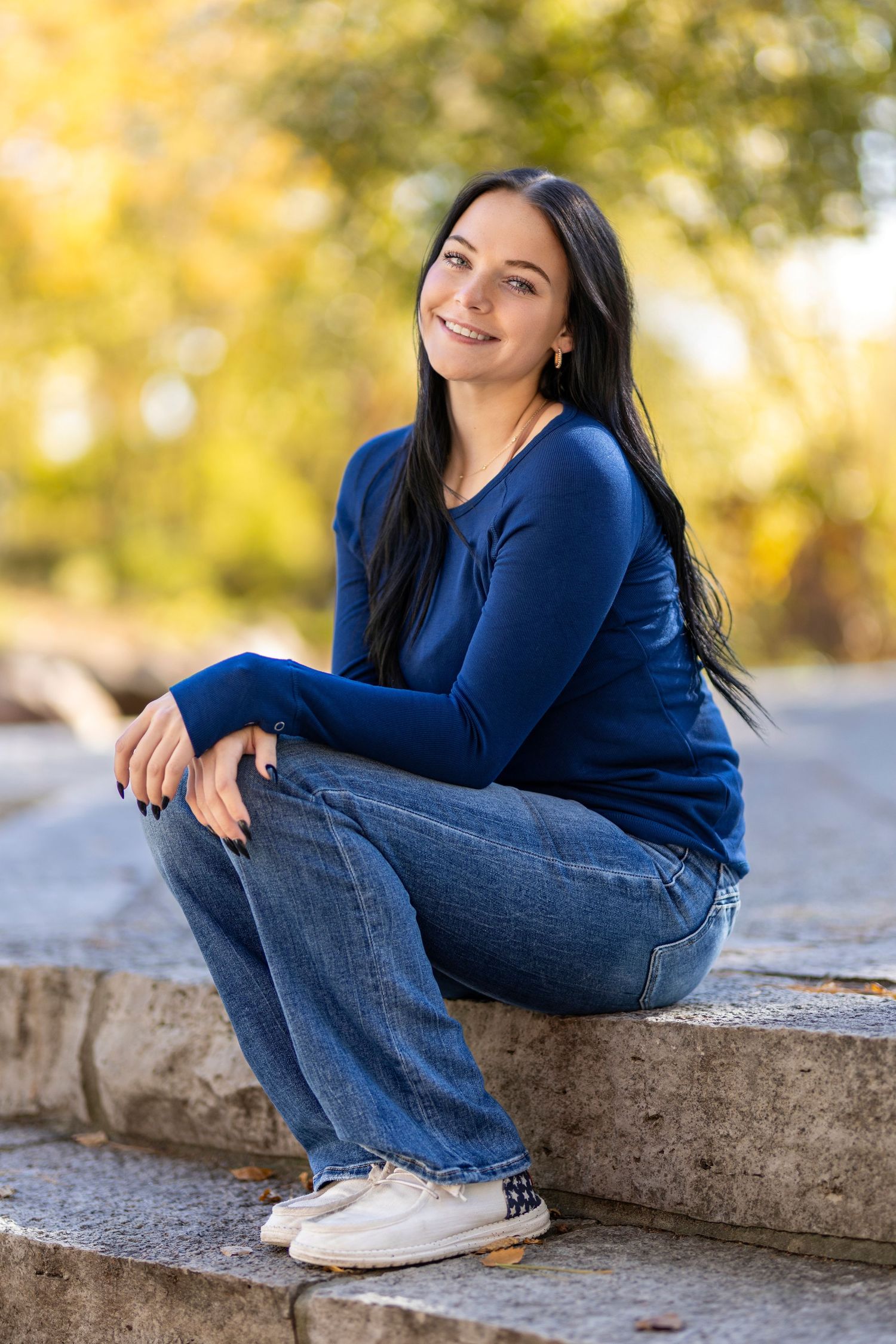 Young woman with dark hair smiling, sitting on stone steps outdoors in blue sweater and jeans on a sunny day.