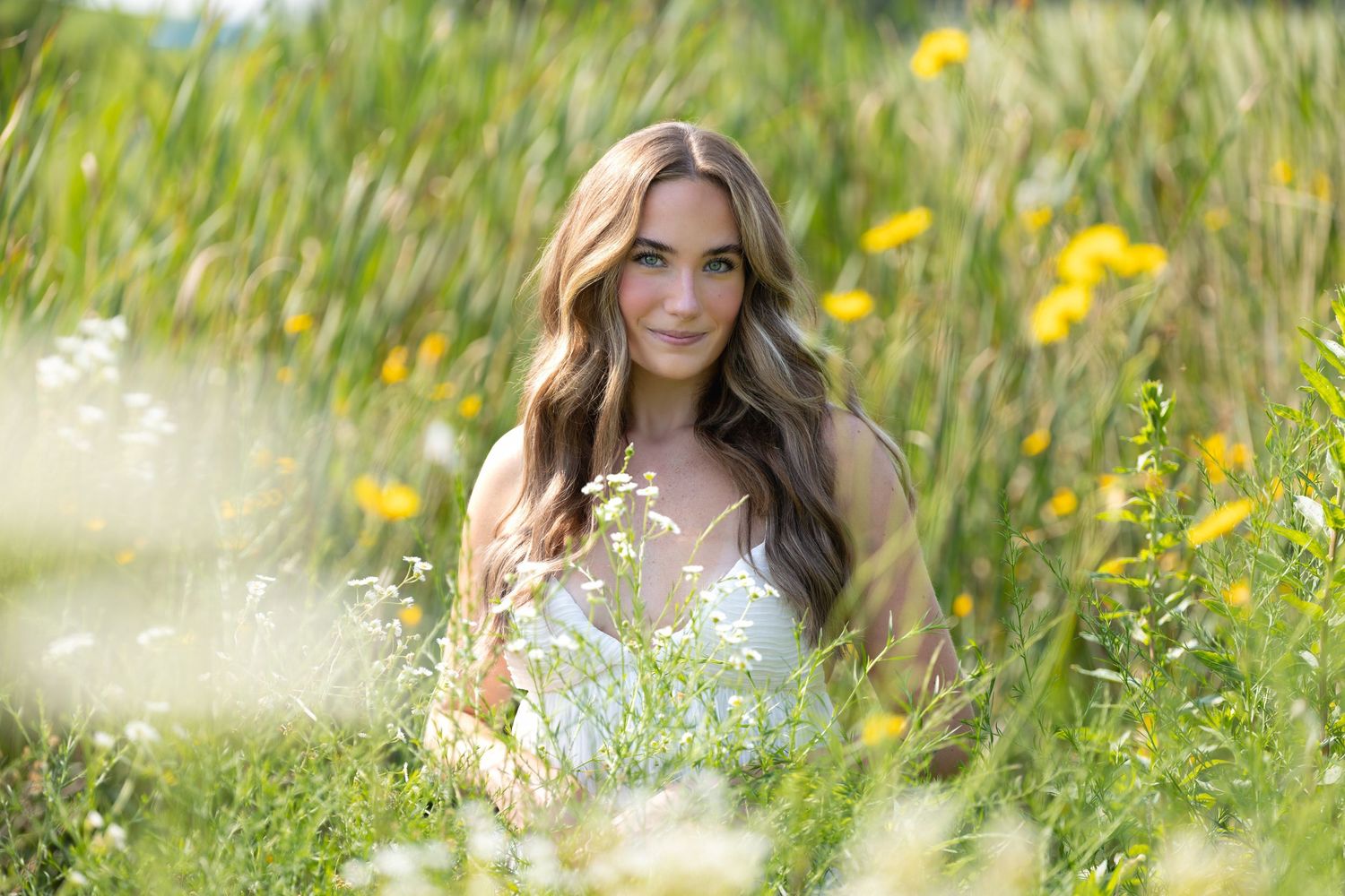 Young woman with long brown hair sits among wildflowers and tall grass in a sunny meadow wearing a white dress.