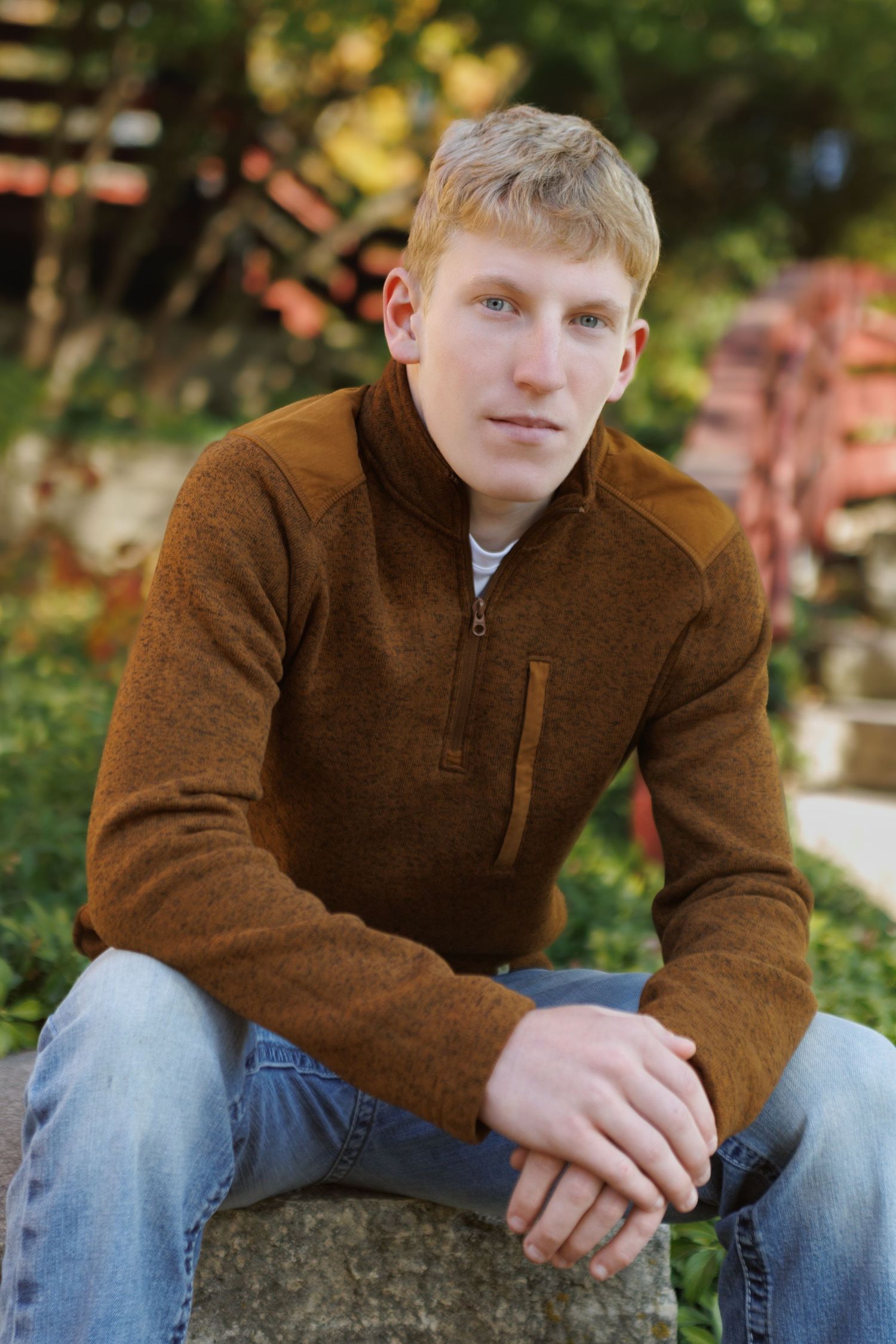 Young man in brown quarter-zip sweater and jeans sitting outdoors on stone, posing for senior portrait photo.