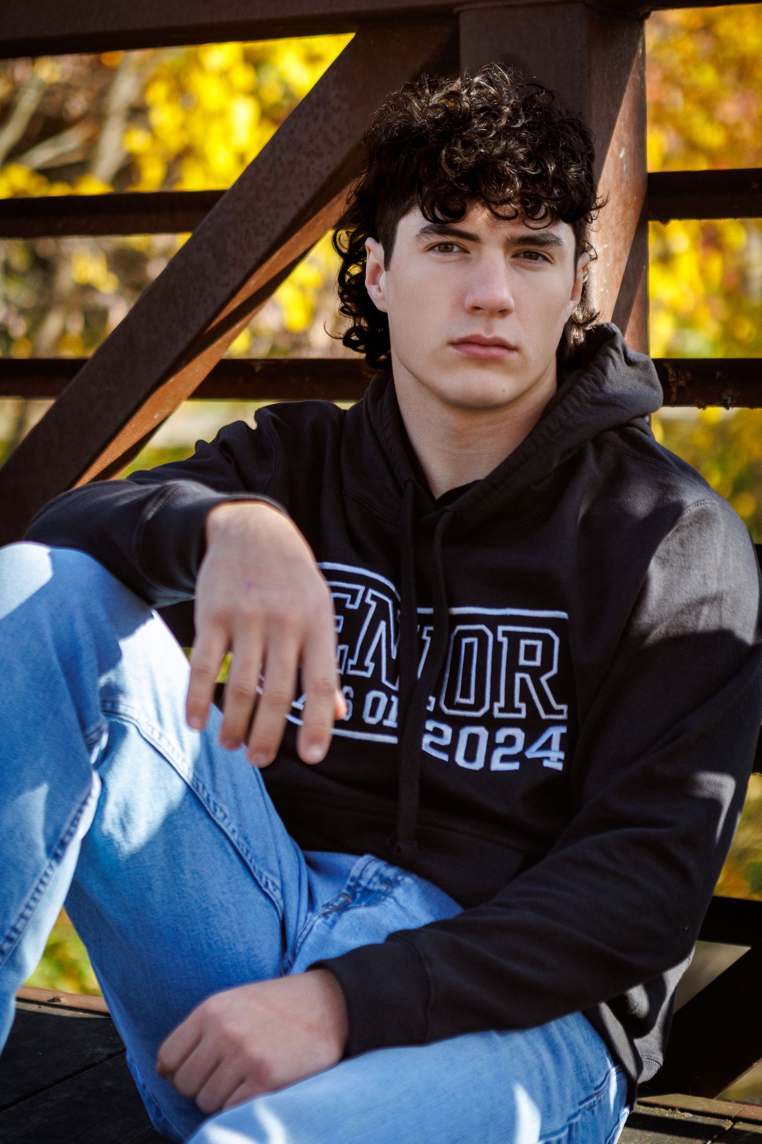 A young man in a black Class of 2024 hoodie and jeans sits casually against a rustic metal fence with autumn foliage.