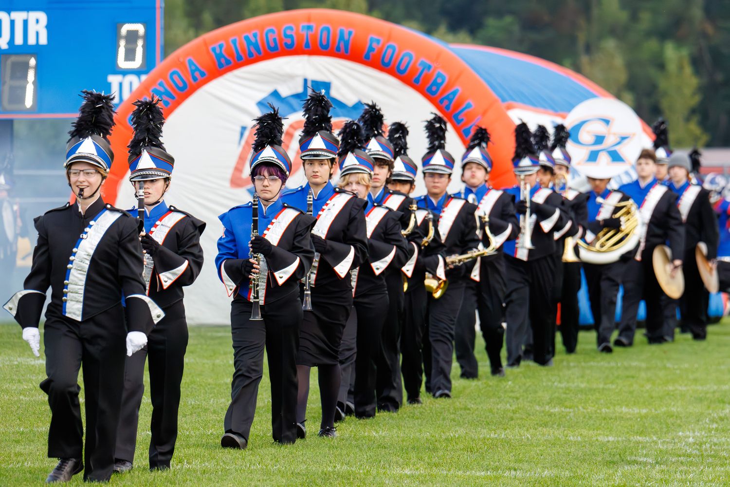 High school marching band in black and white uniforms performs in formation on a sports field under a red arch.