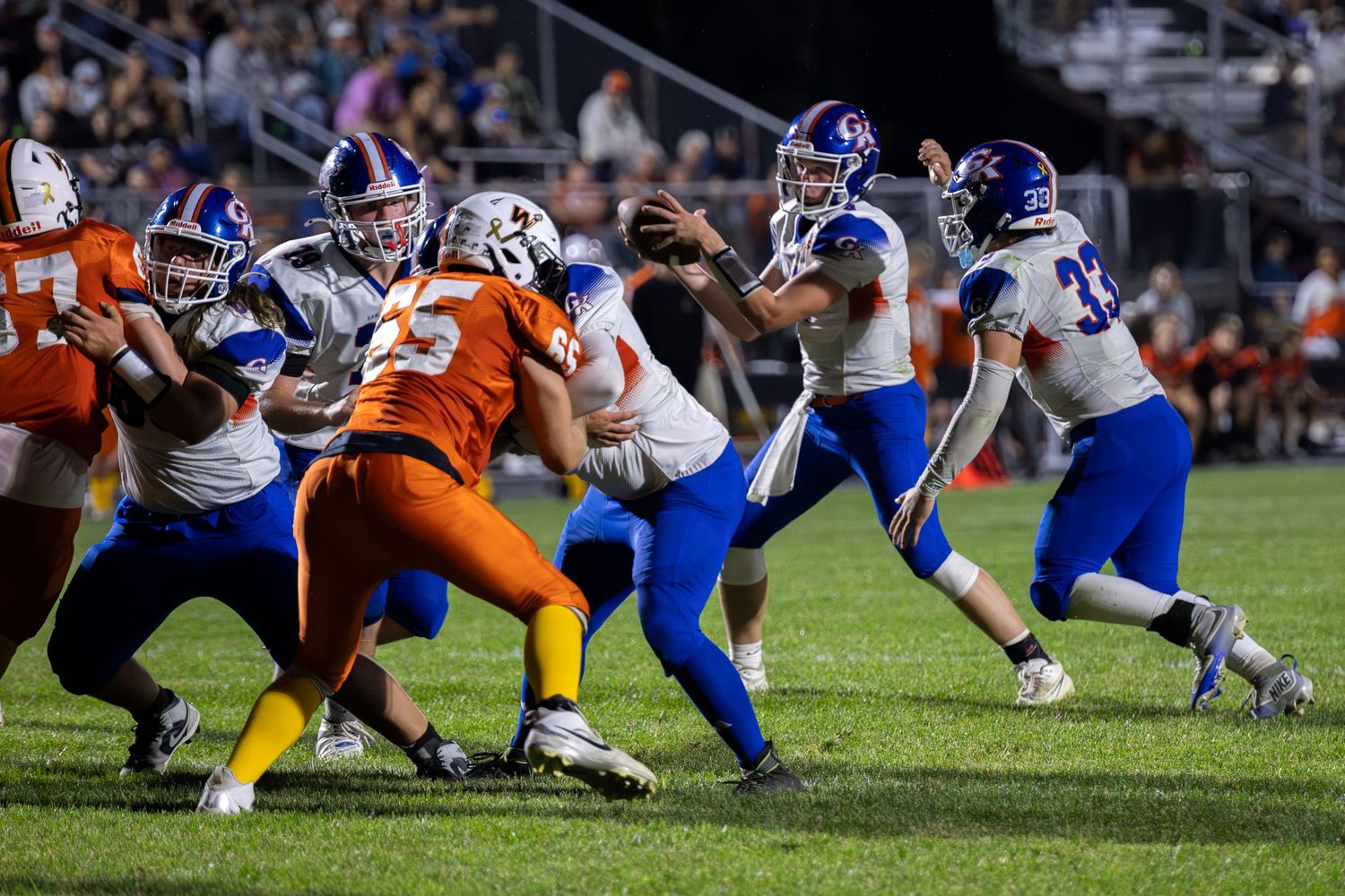 High school football players in blue and orange uniforms compete during an intense game under stadium lights.