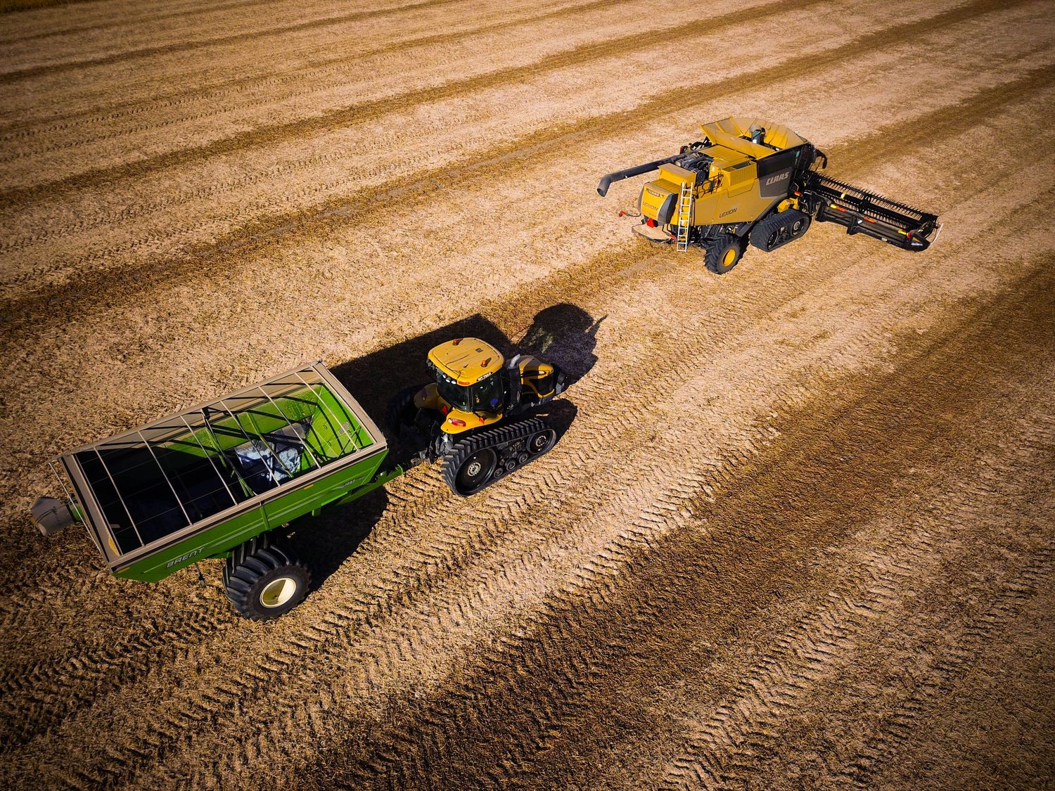Aerial view of a yellow combine harvester unloading grain into a green trailer in a harvested wheat field.