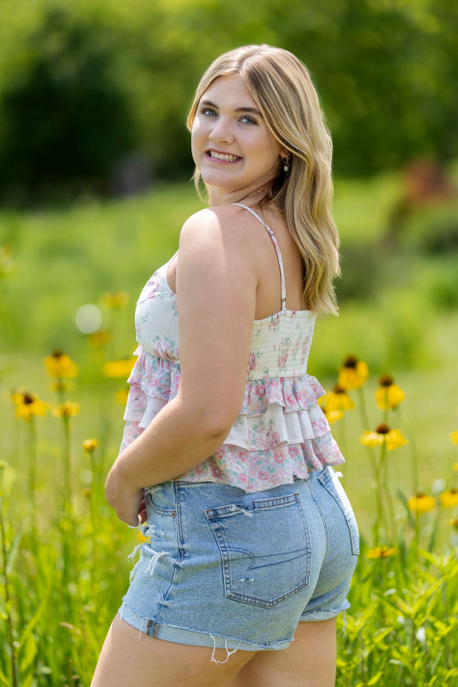 Young blonde woman in floral top and denim shorts smiling in a sunny field with yellow wildflowers.