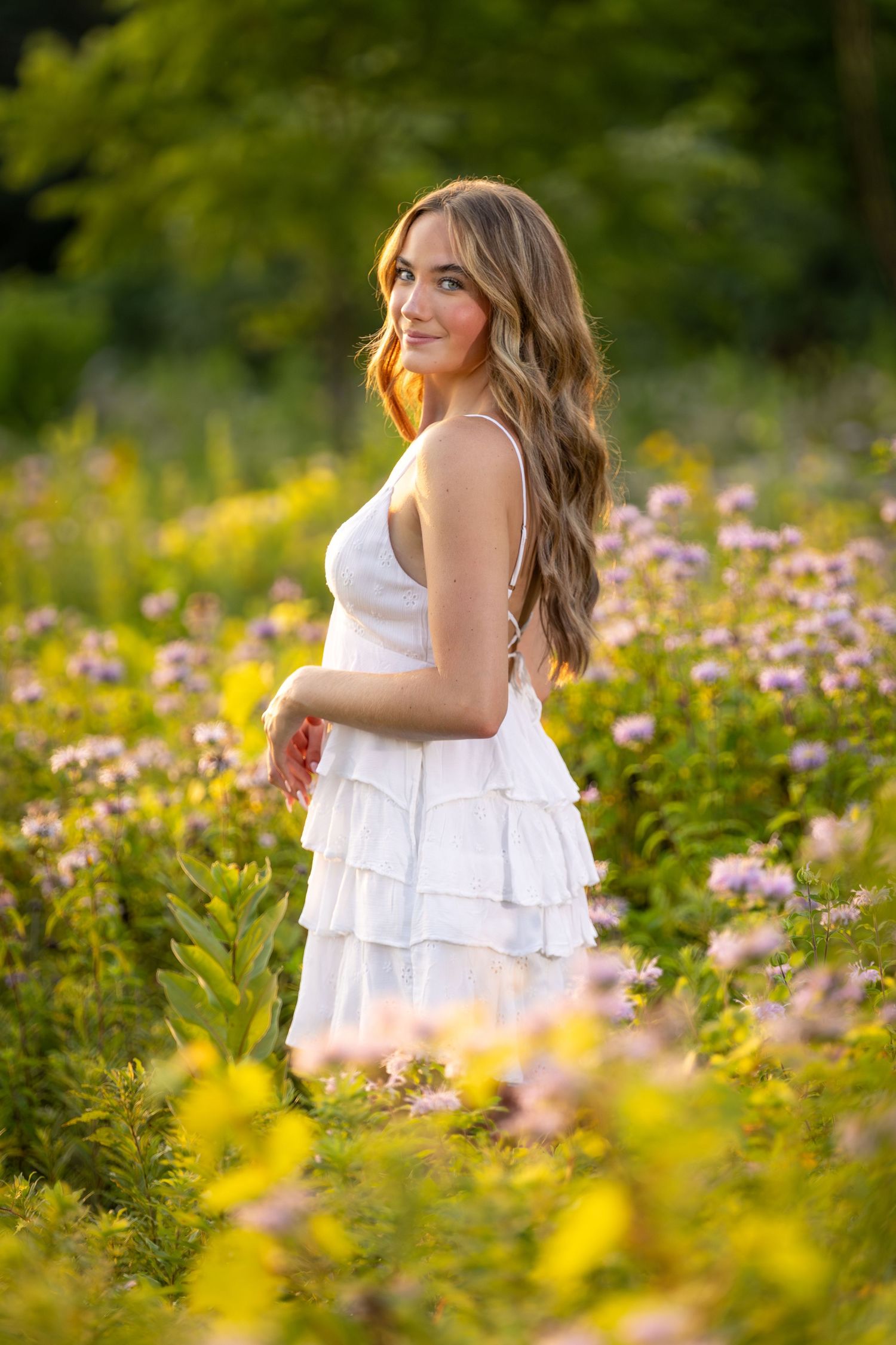 Young woman in white ruffled dress standing in wildflower field with purple and yellow blooms at golden hour.