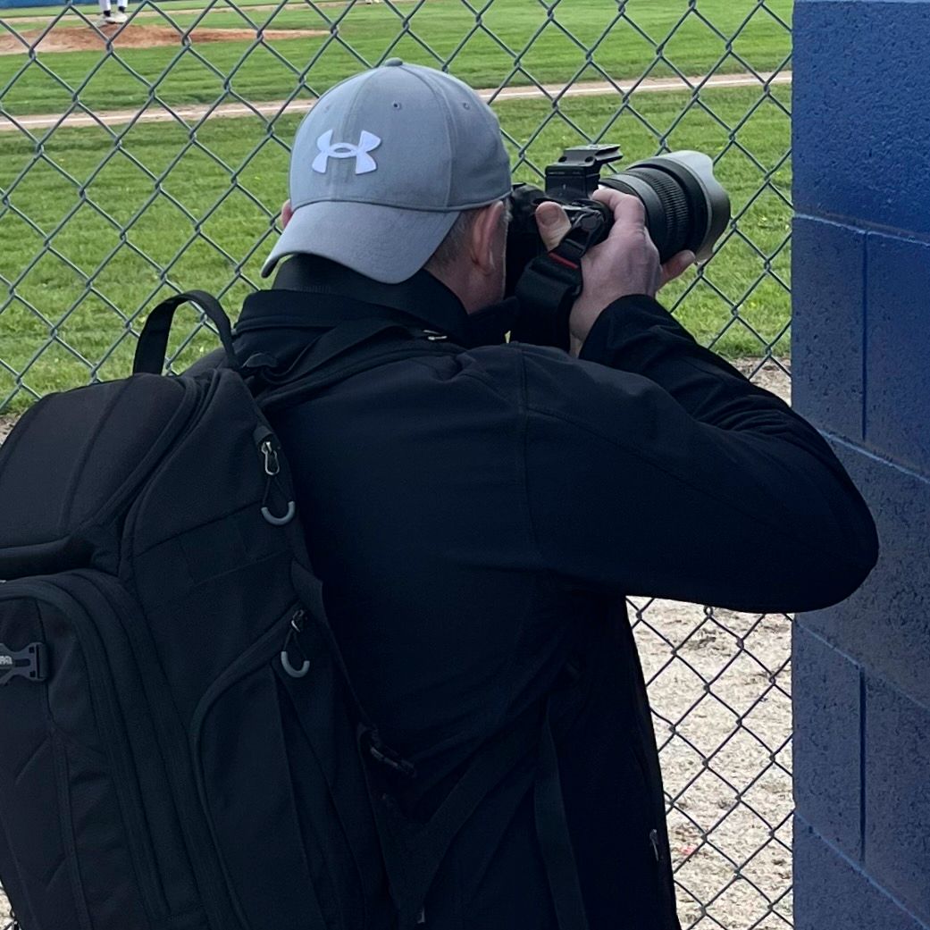 Photographer wearing Under Armour cap and black backpack shooting with DSLR camera at a baseball field.