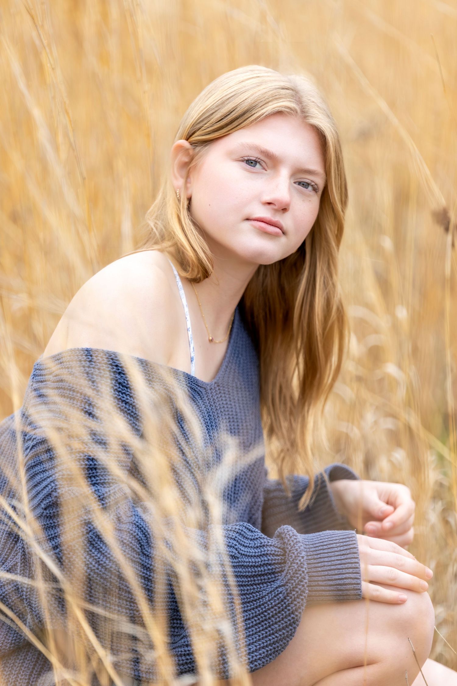 A teenage girl with long blonde hair sits in golden tall grass, wearing a blue knit sweater with an off-shoulder style.