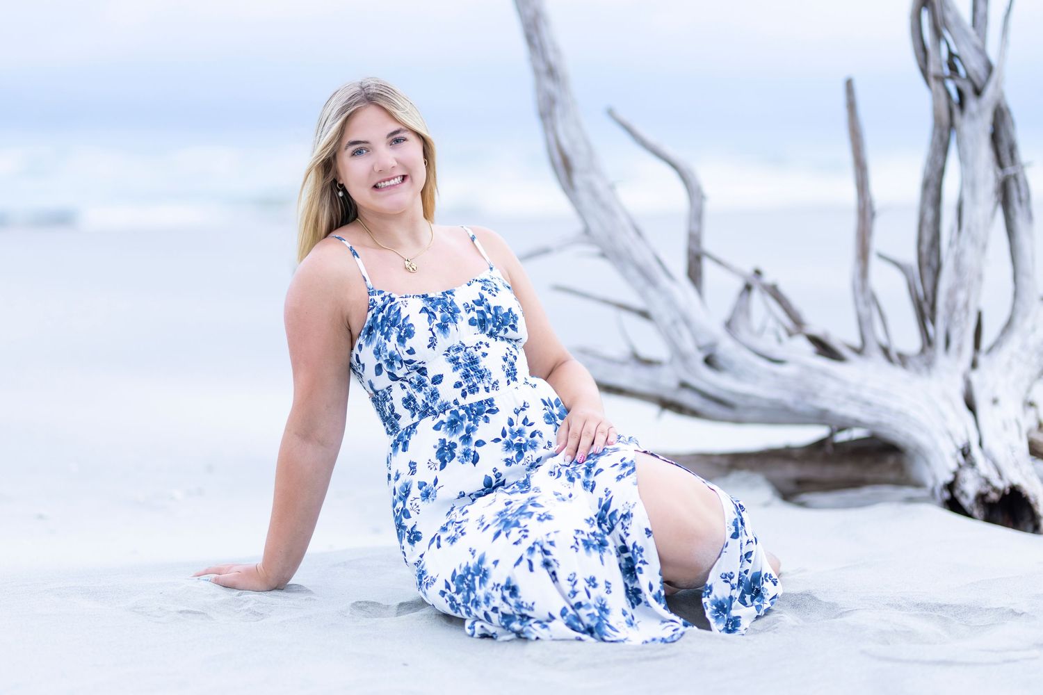 Young blonde woman in blue floral maxi dress posing on sandy beach with driftwood, smiling outdoors.