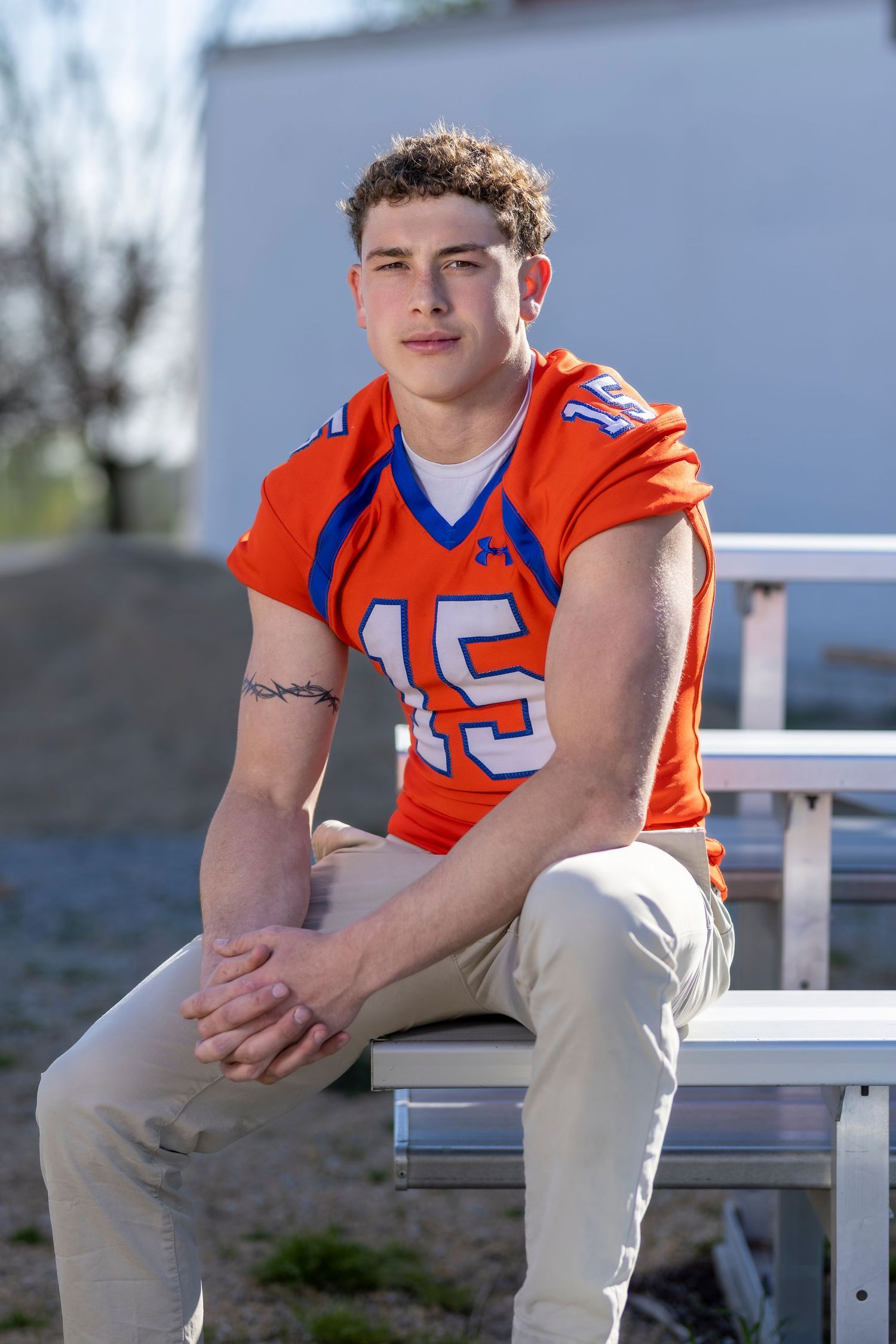 Young male athlete in orange and blue Under Armour football jersey number 15 sitting on bleachers outdoors.