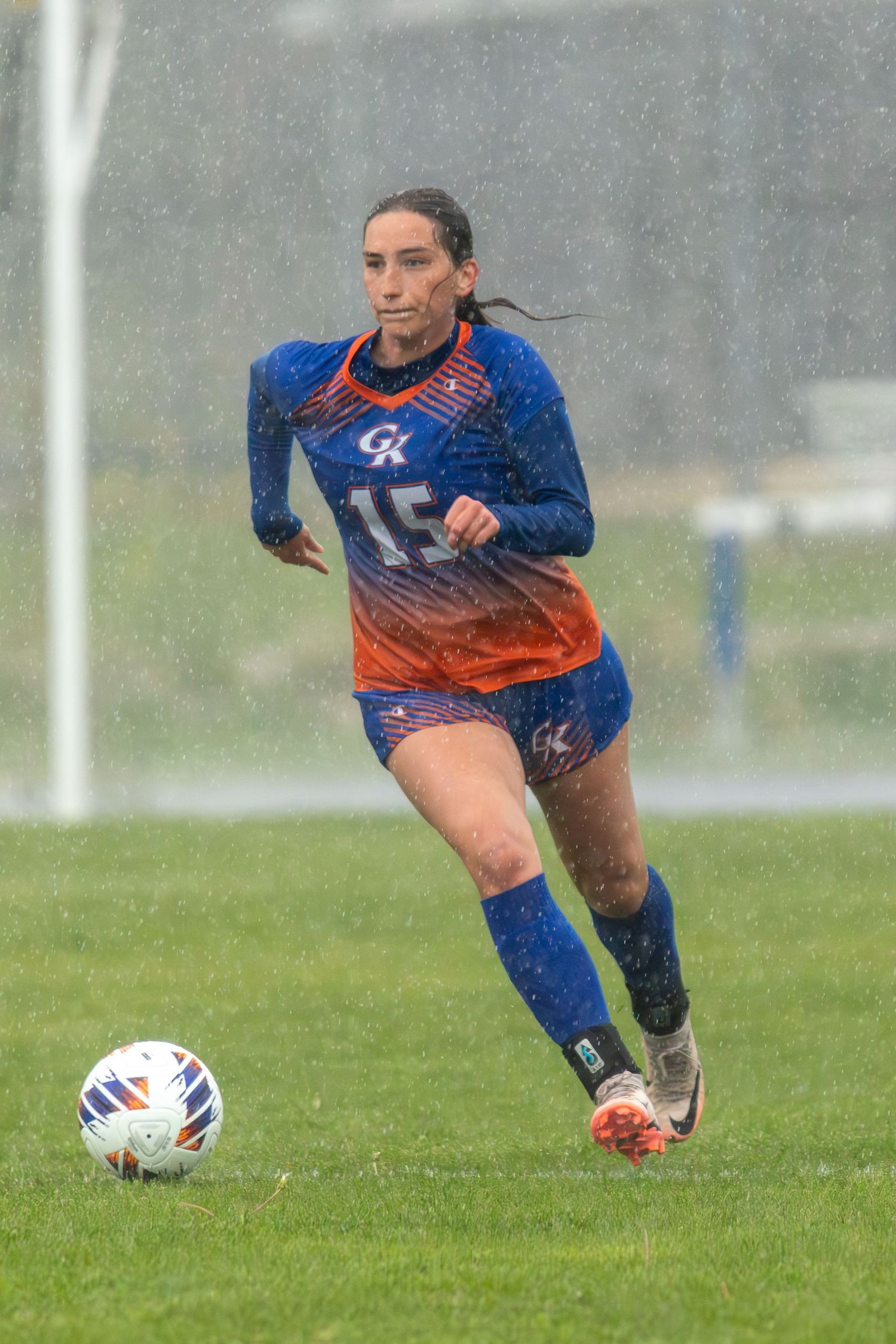 Female soccer player #15 in blue and orange jersey dribbles ball in heavy rain during outdoor match.