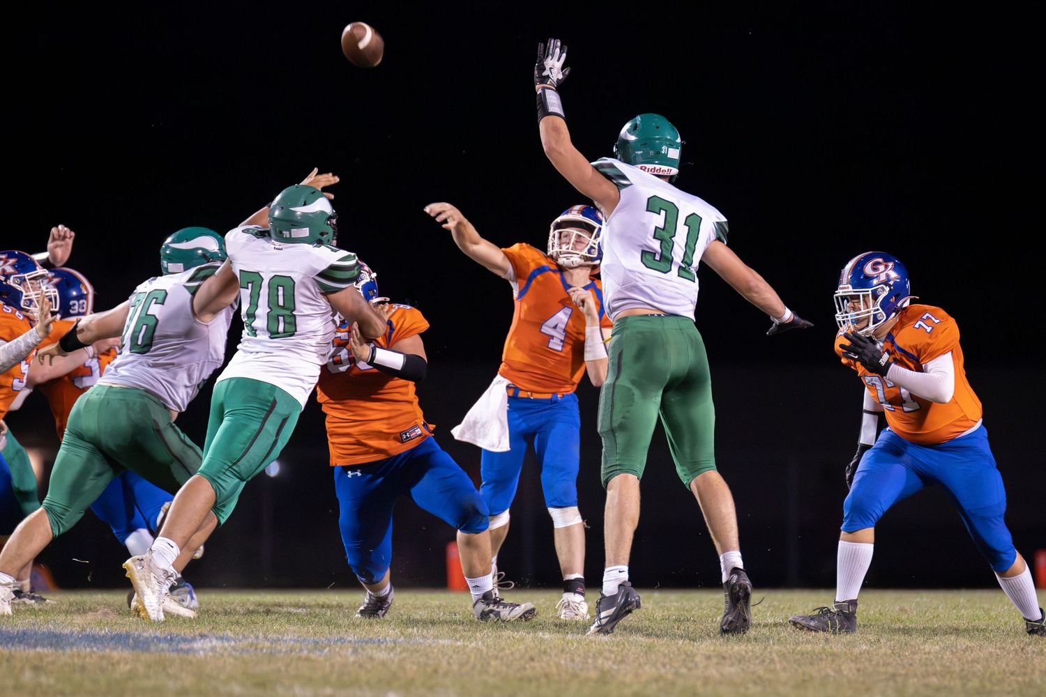 Football player in blue jersey throws a pass during a night game as green-jersied defenders attempt to block.