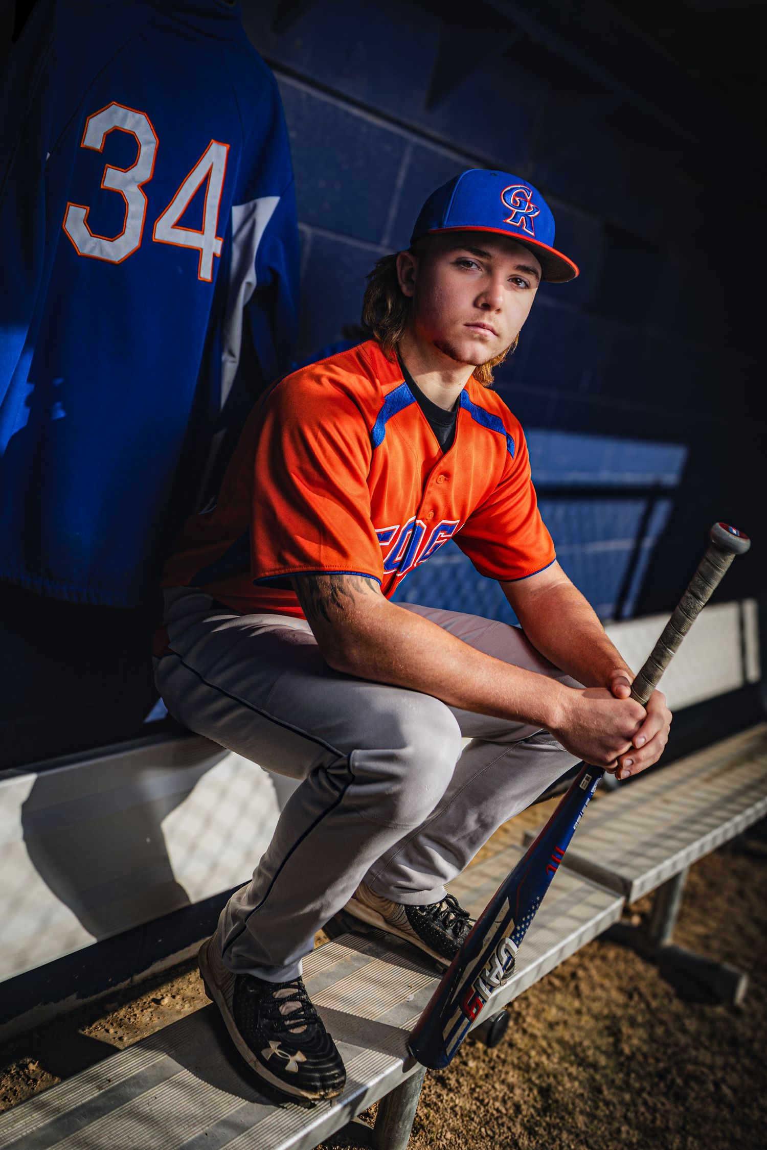 Baseball player in orange uniform sits in dugout with bat during game.