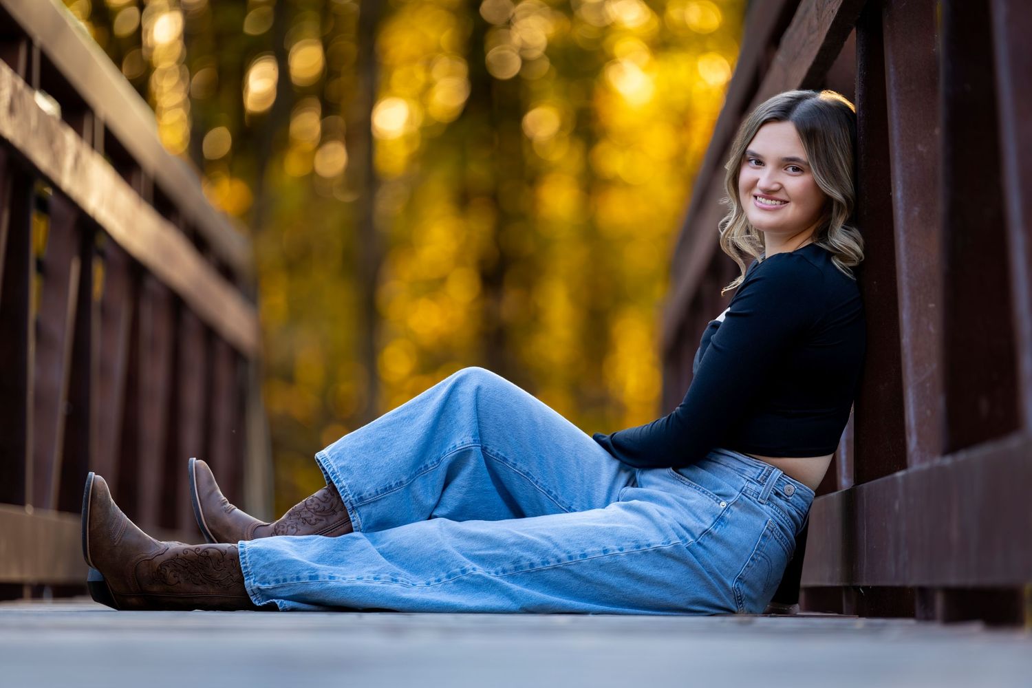 Young woman in jeans and boots sitting on a wooden bridge, smiling, with golden autumn trees in background.