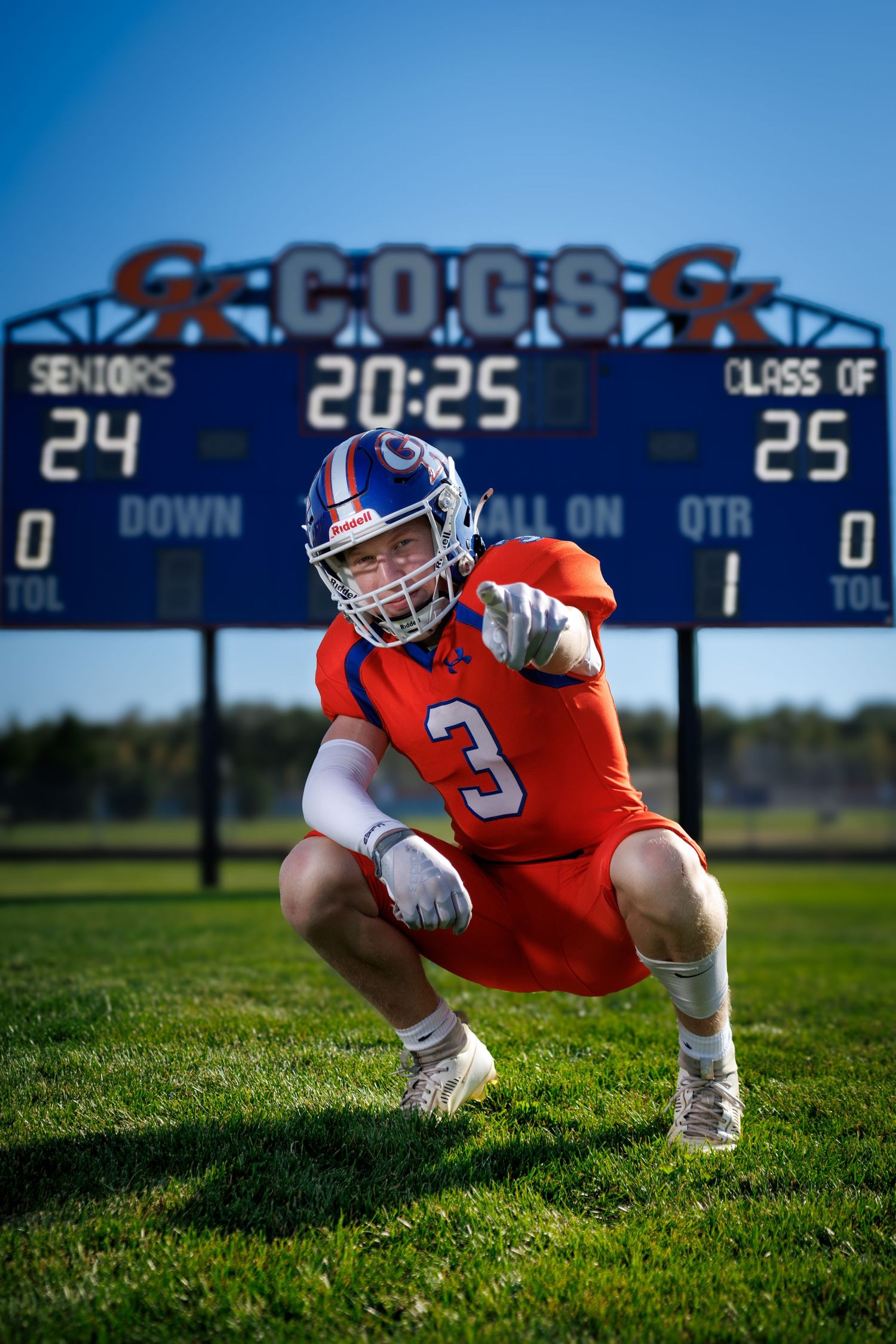 Football player #3 in orange uniform crouches in ready stance on field with Cogs scoreboard behind him.