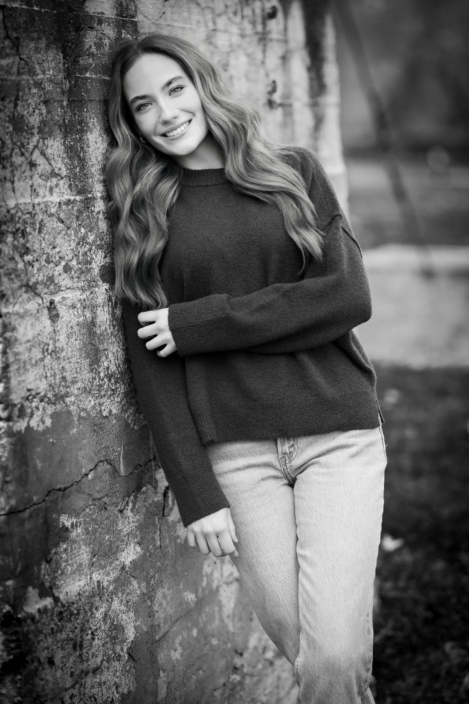 Young woman with wavy hair leaning against stone wall in black and white portrait.