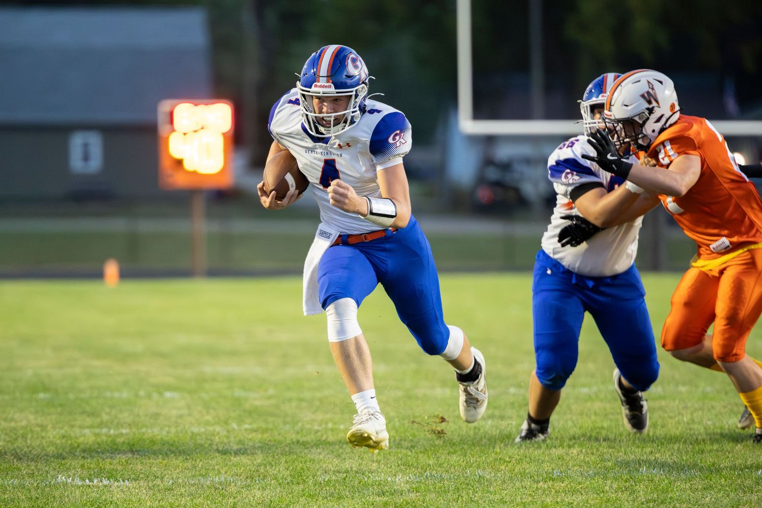 High school football player in blue and white uniform runs with ball during night game against orange-uniformed opponents.